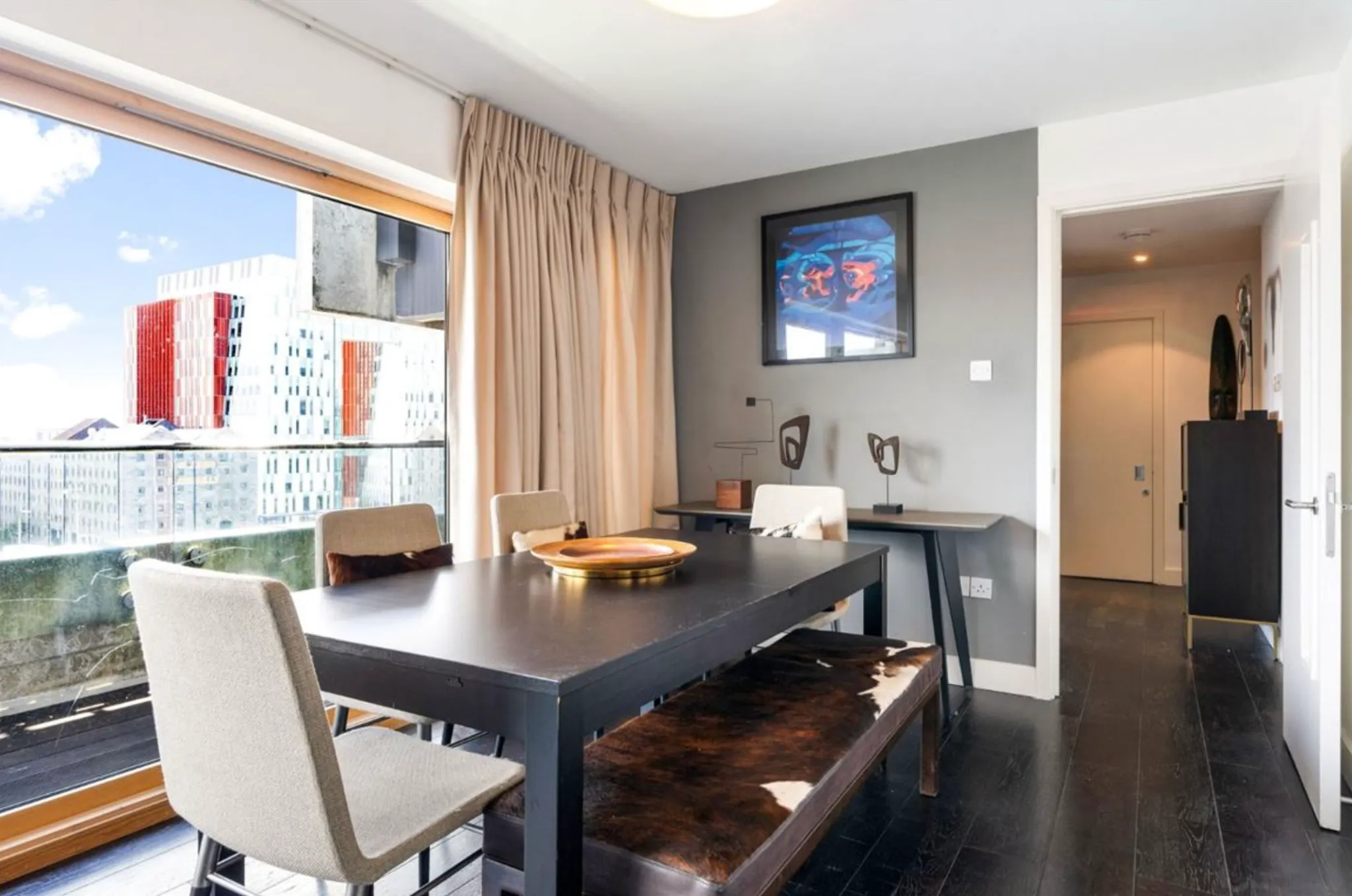Dining room with a dark rectangular table, upholstered chairs and a bench, large floor-to-ceiling window with curtains and a framed artwork on a gray wall.