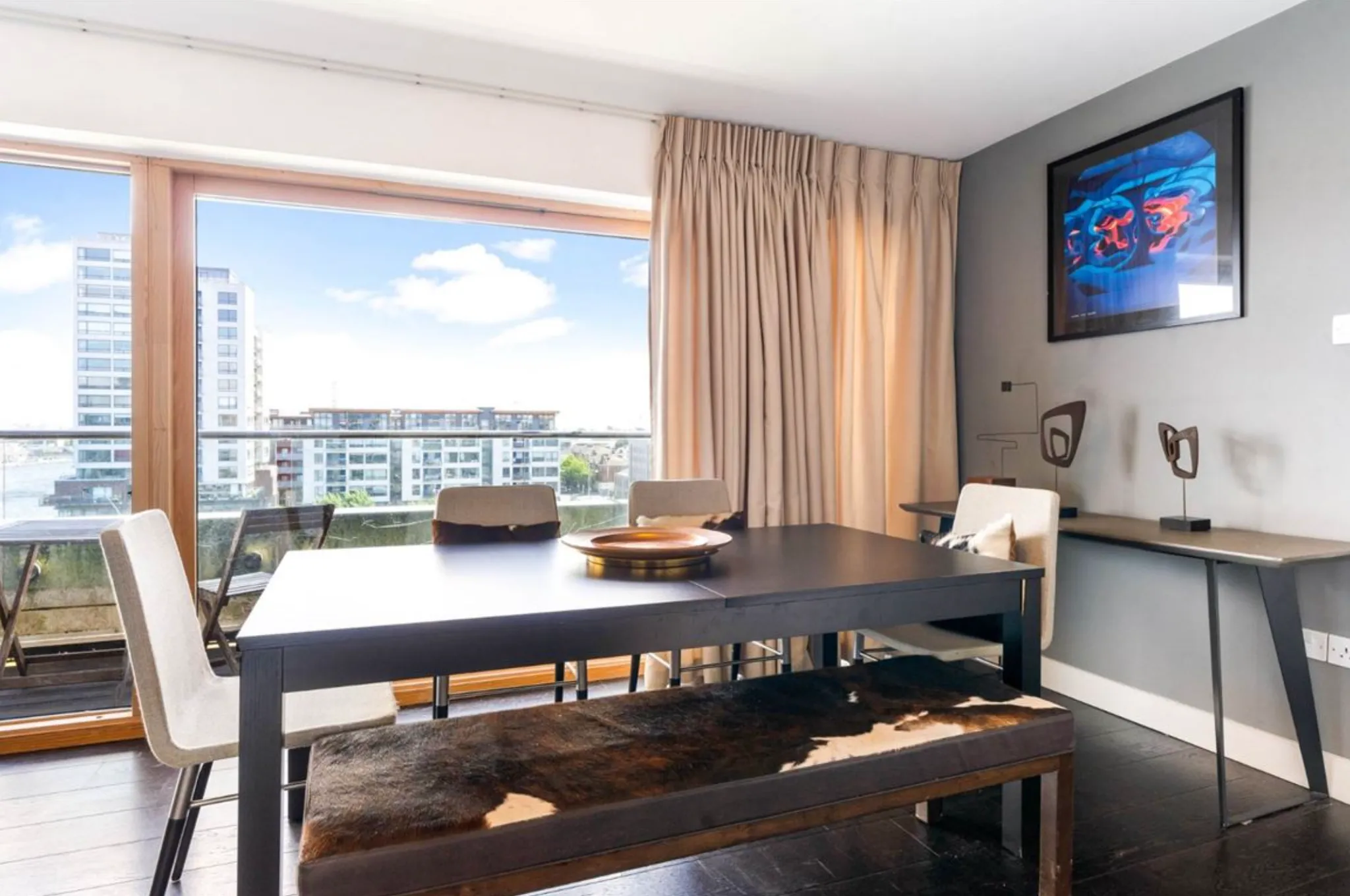Dining room with a dark rectangular table, upholstered chairs, a bench with a fur cover, large floor-to-ceiling window and curtains, framed wall art and a side desk.