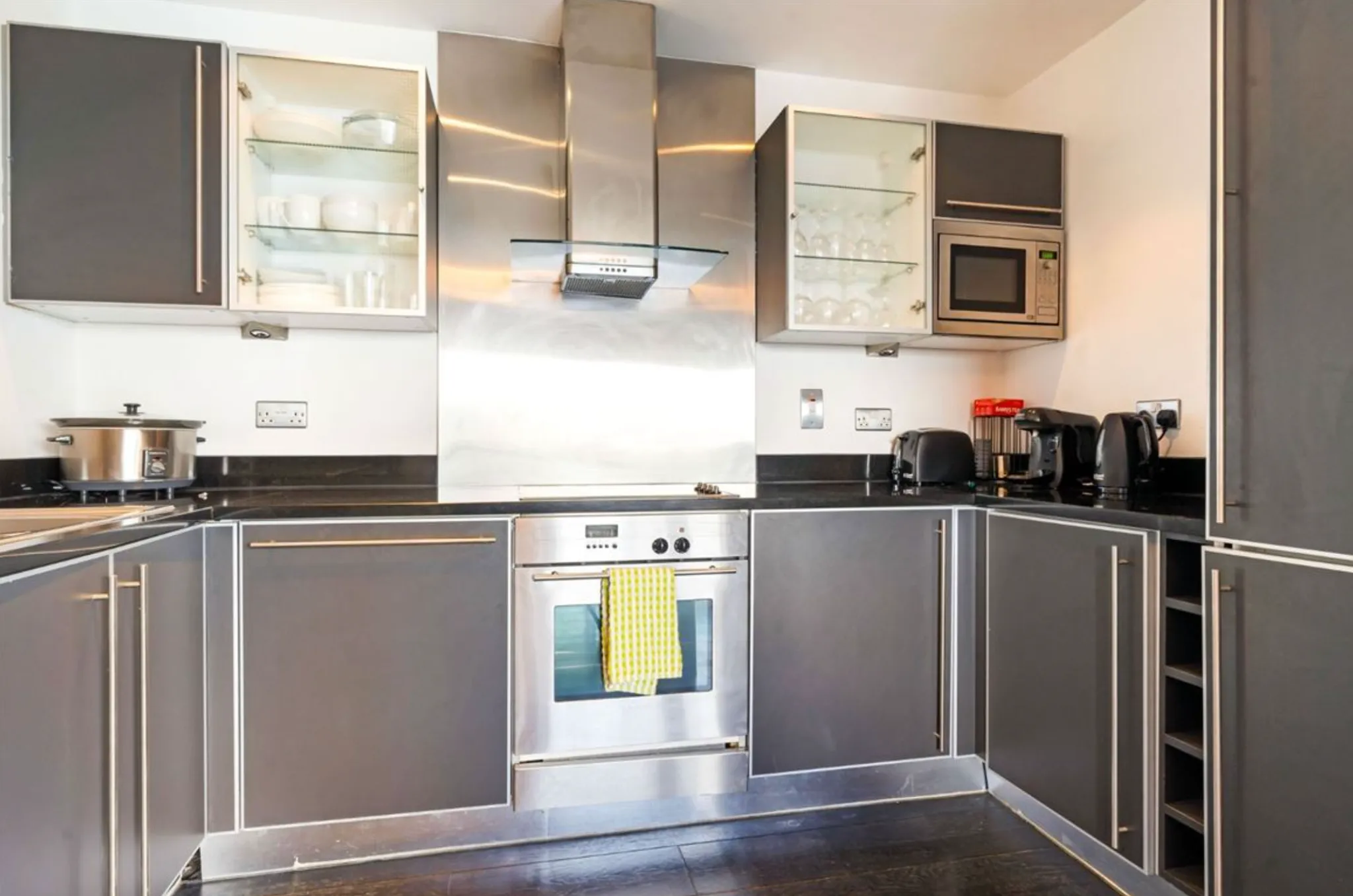 Kitchen with stainless steel oven and range hood, dark cabinets, glass-front upper cabinets, microwave, and small countertop appliances.