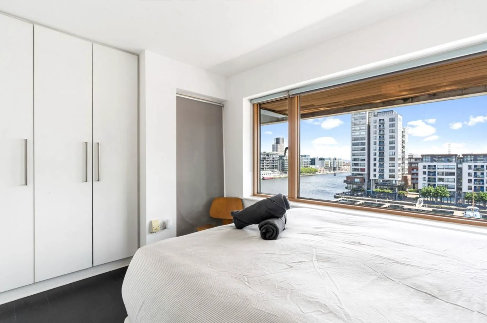 Bedroom with a double bed, built-in white wardrobe, chair, and large window showing a waterfront city view.