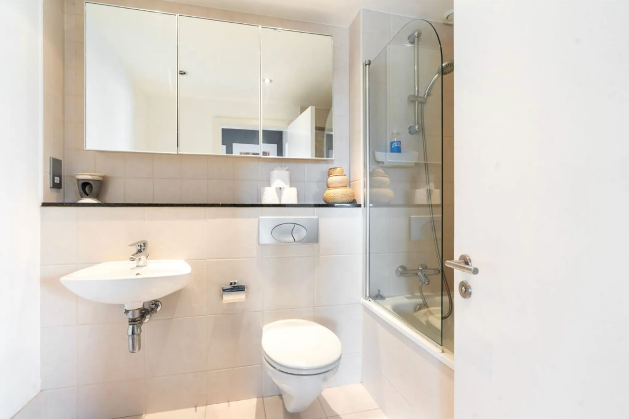 Bathroom with wall-mounted sink, wall-mounted toilet, mirrored medicine cabinet and a bathtub with glass shower screen and handheld showerhead.