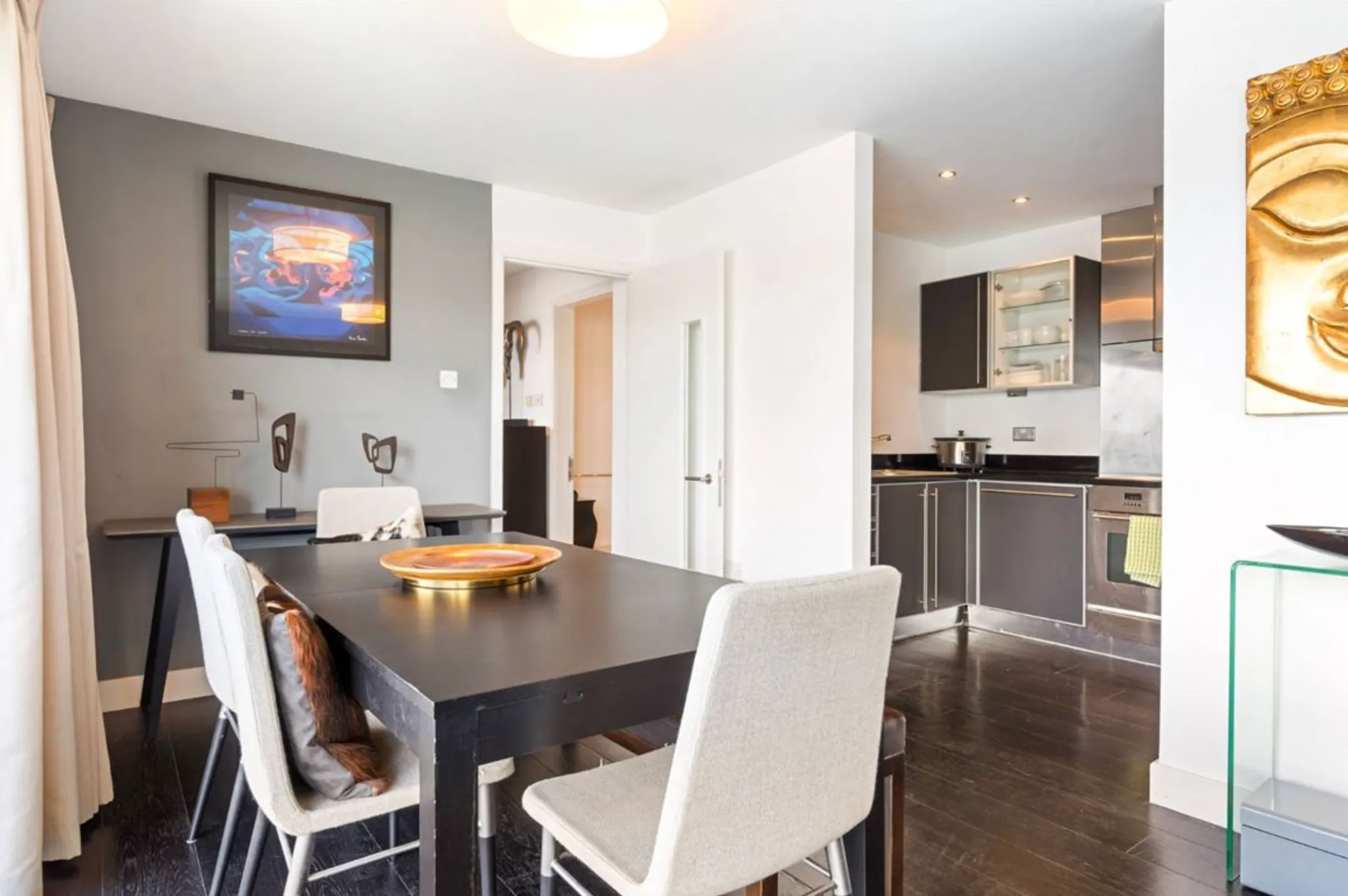 Dining area with a dark rectangular table, upholstered chairs, wall art, and an open-plan kitchen visible to the right.