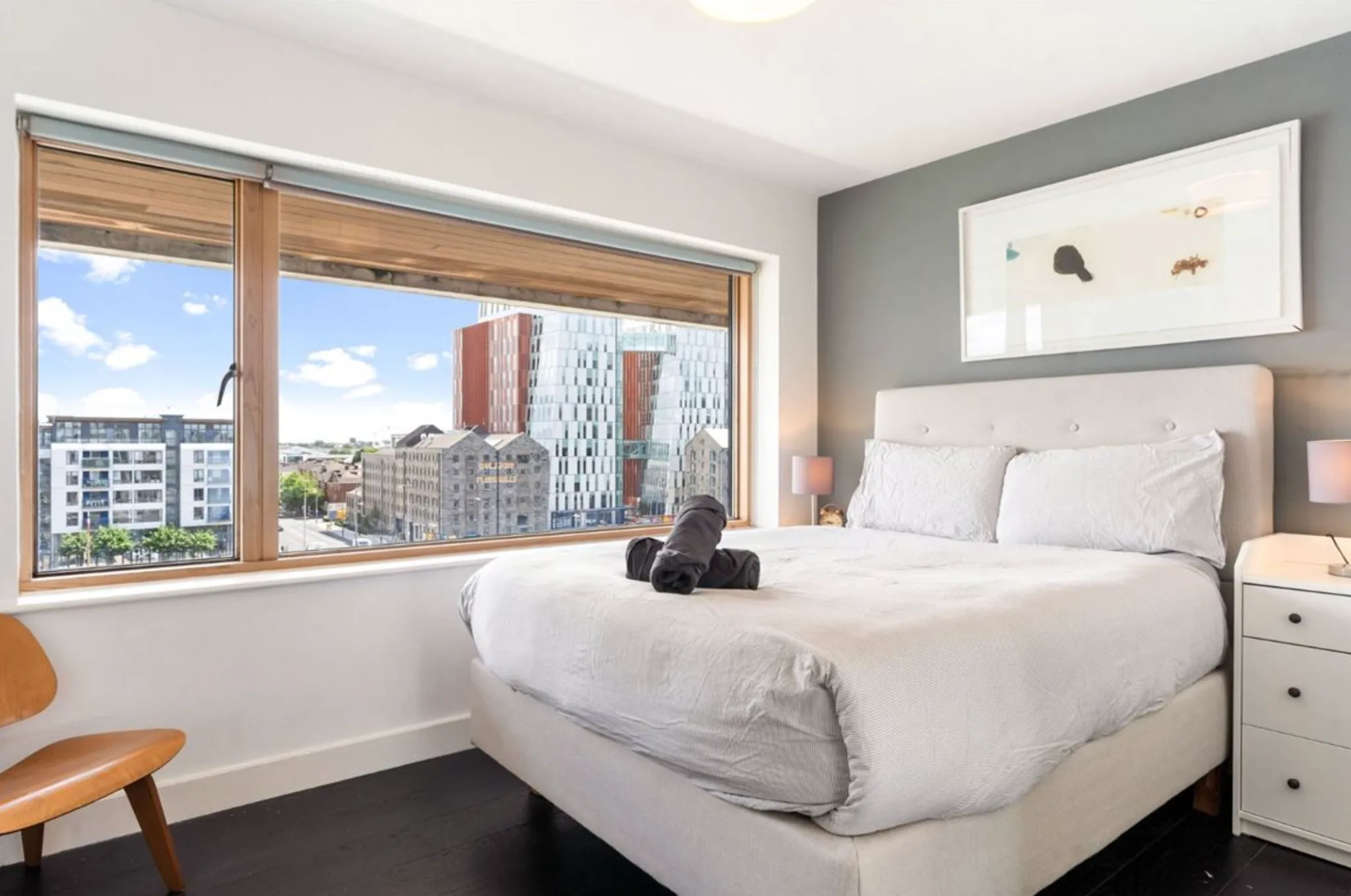 Bedroom with a double bed, upholstered headboard, large window showing a city view, bedside table with lamp, framed artwork, and a chair.