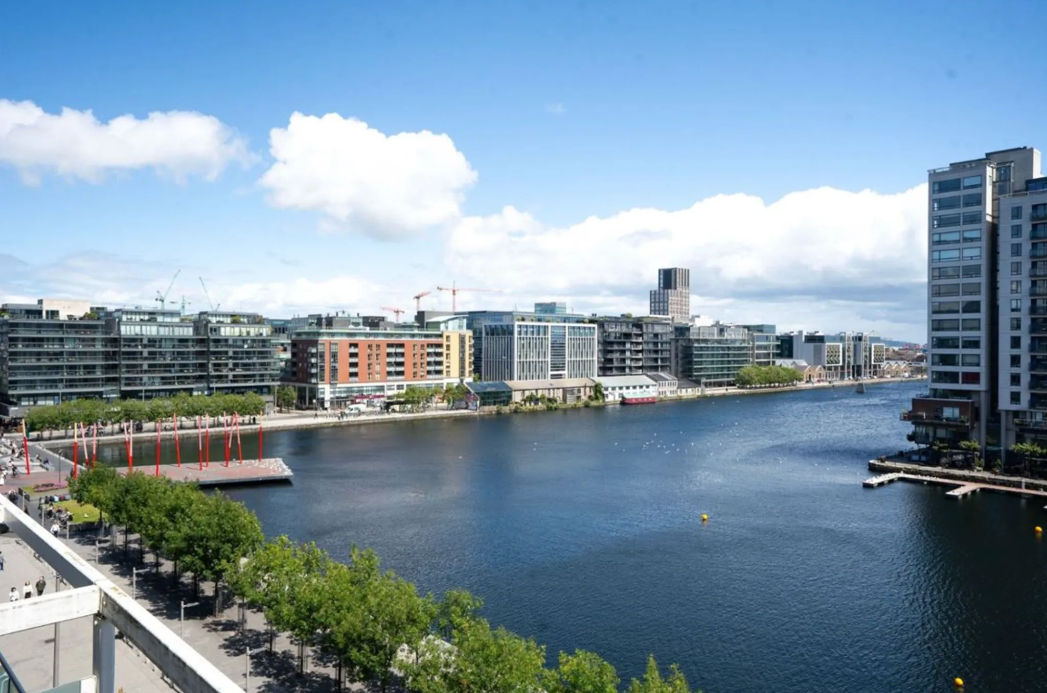 Waterfront city view showing a river or dock, low-rise and high-rise buildings along the opposite bank, a promenade with trees, and a railing in the foreground.