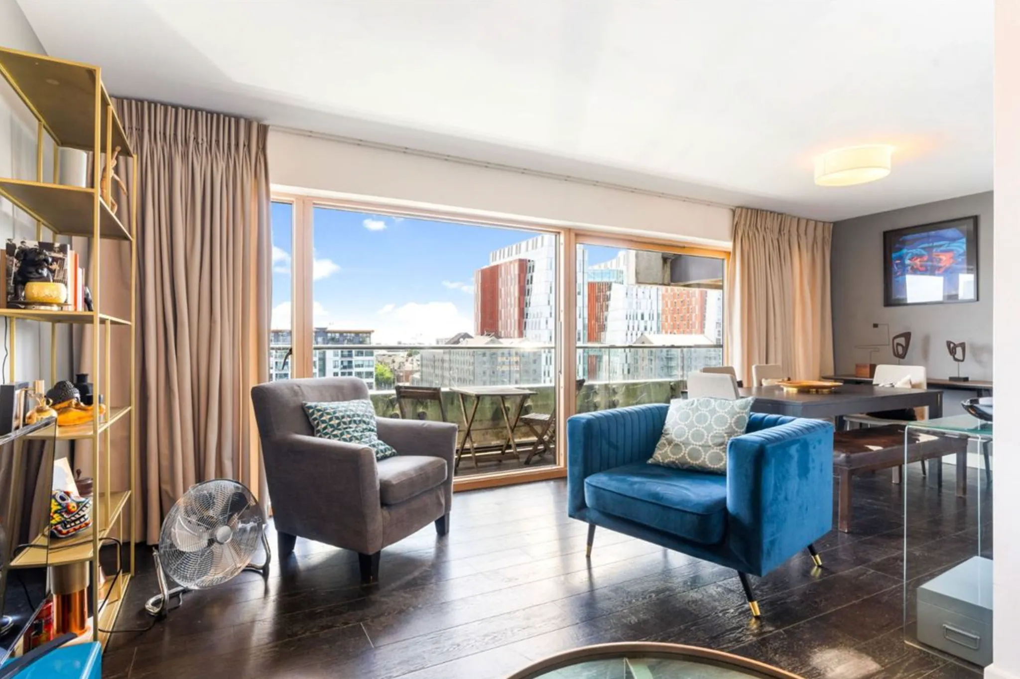 Living room with two armchairs and cushions, a dining table, bookshelf, and large sliding glass door opening to a balcony with city view.