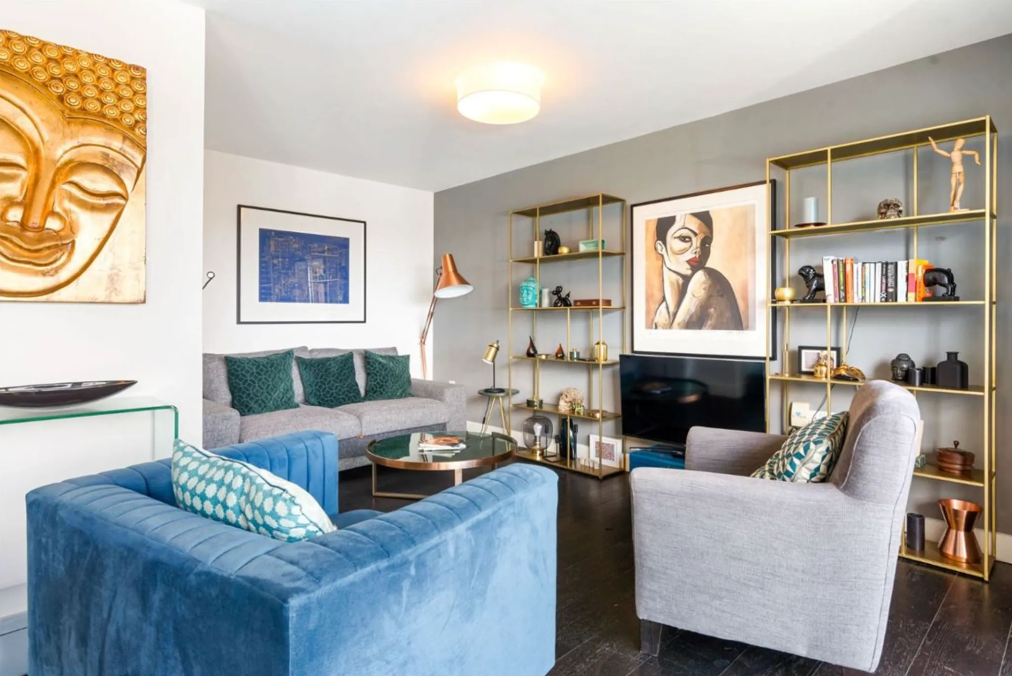 Living room with a blue upholstered sofa, grey armchair, round glass coffee table, gold open shelving unit, TV and framed wall art.