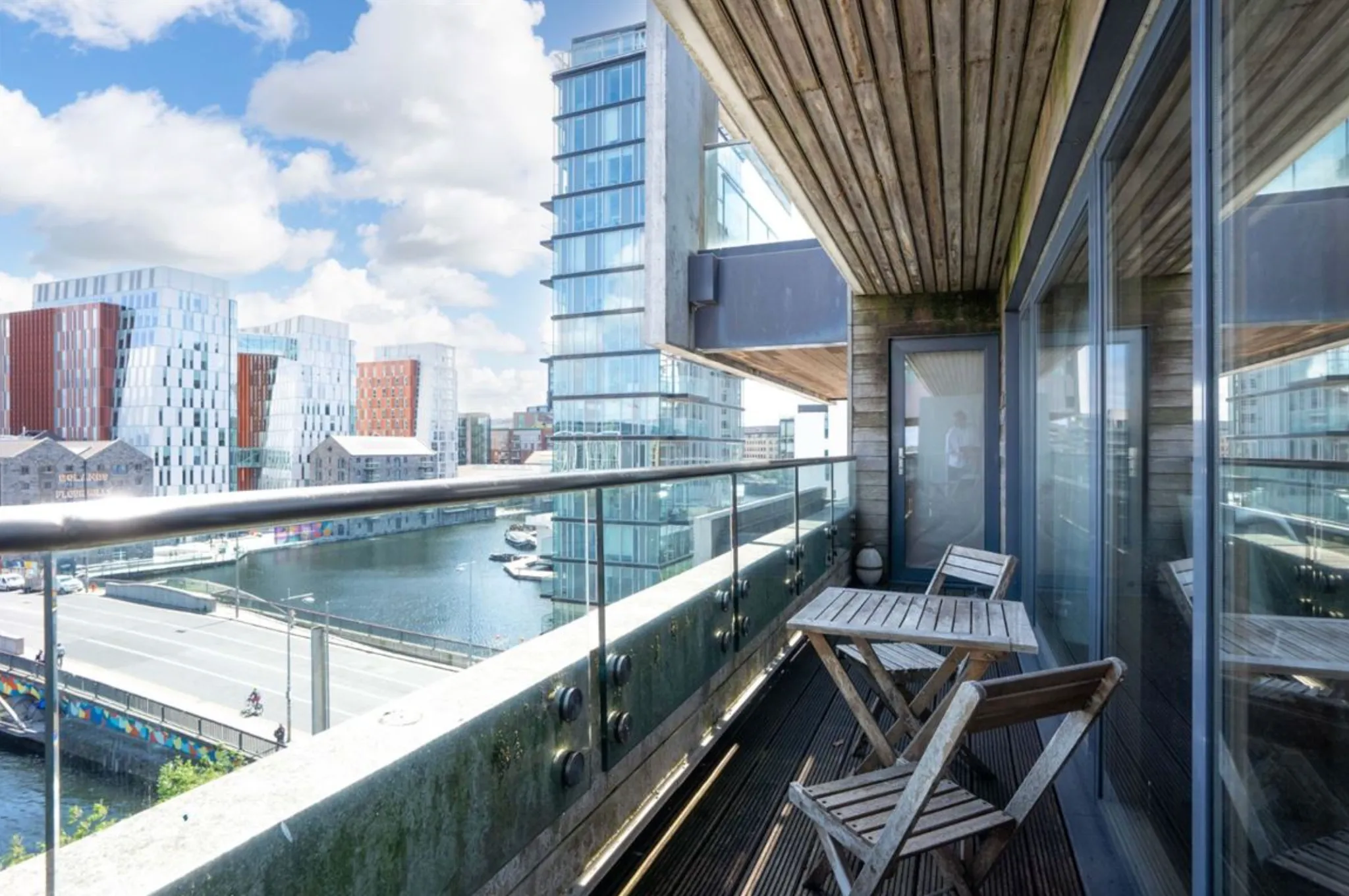 Balcony with glass railing, two wooden folding chairs and a small table overlooking a waterfront and modern buildings