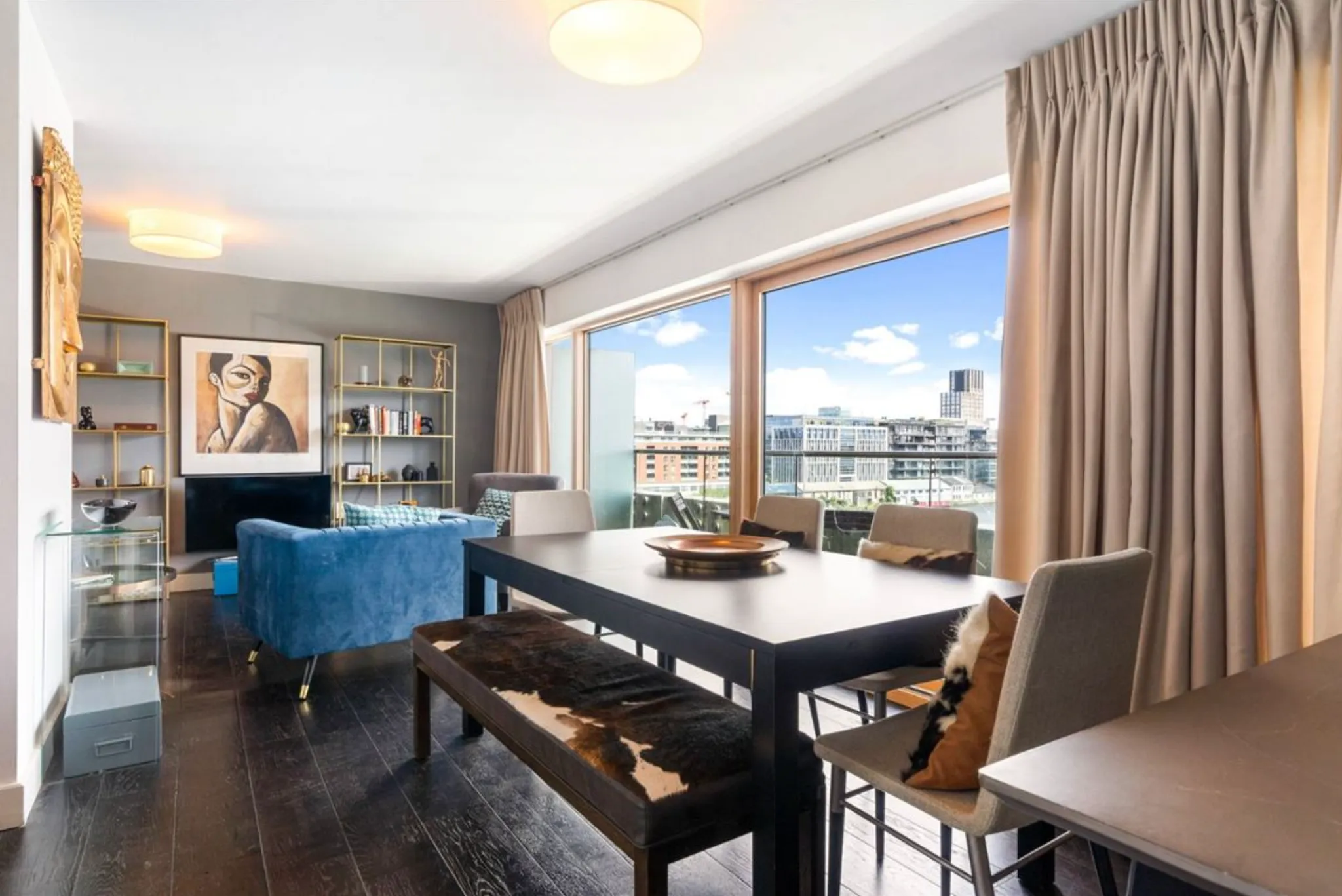 Open-plan living and dining area with a dining table and chairs, bench, blue sofa, shelving, artwork on the wall and large floor-to-ceiling windows with curtains showing a city view.