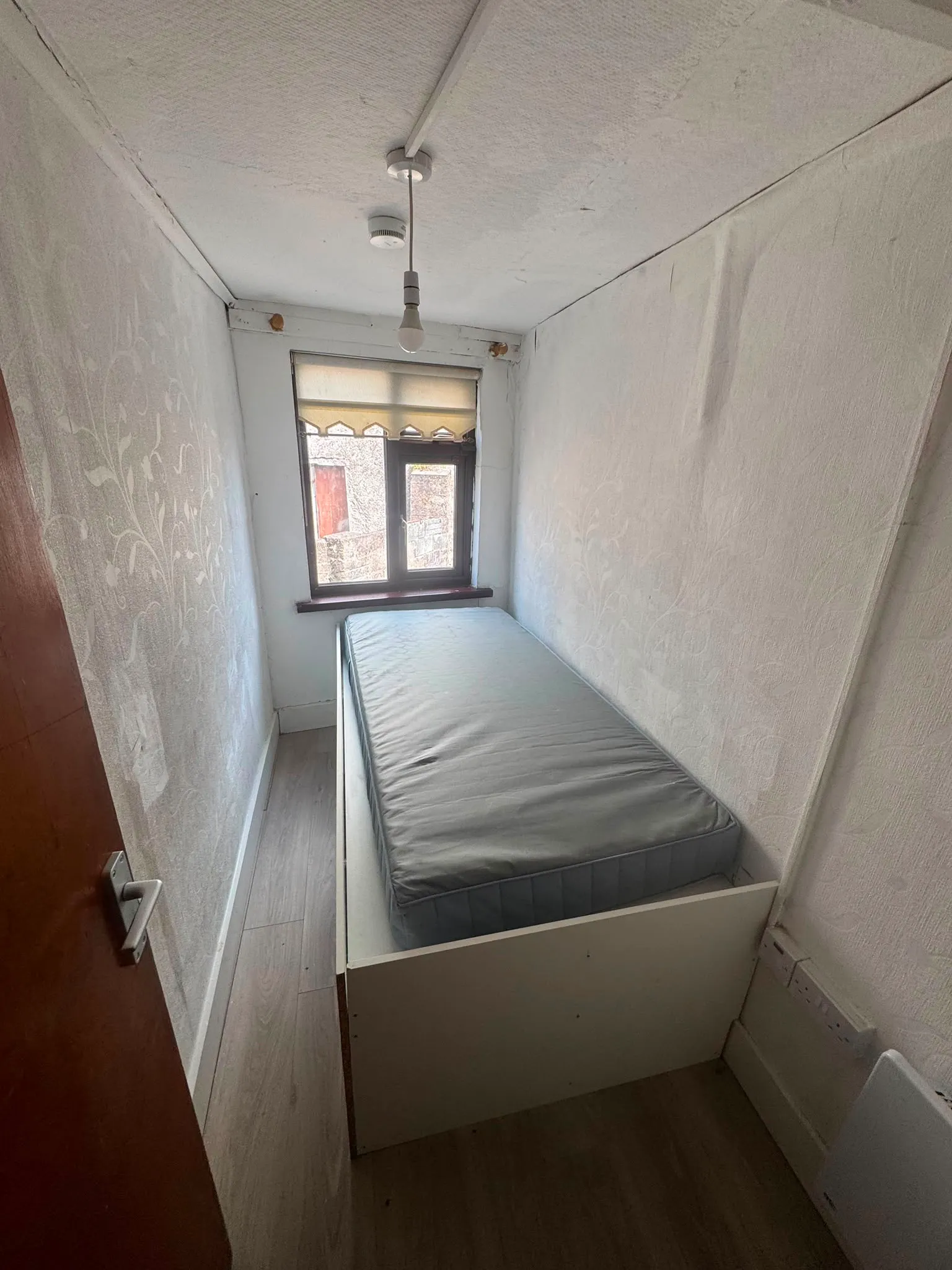 Narrow bedroom with a single bed placed along the wall, a window with a roller blind at the far end, and a hanging lightbulb from the ceiling.