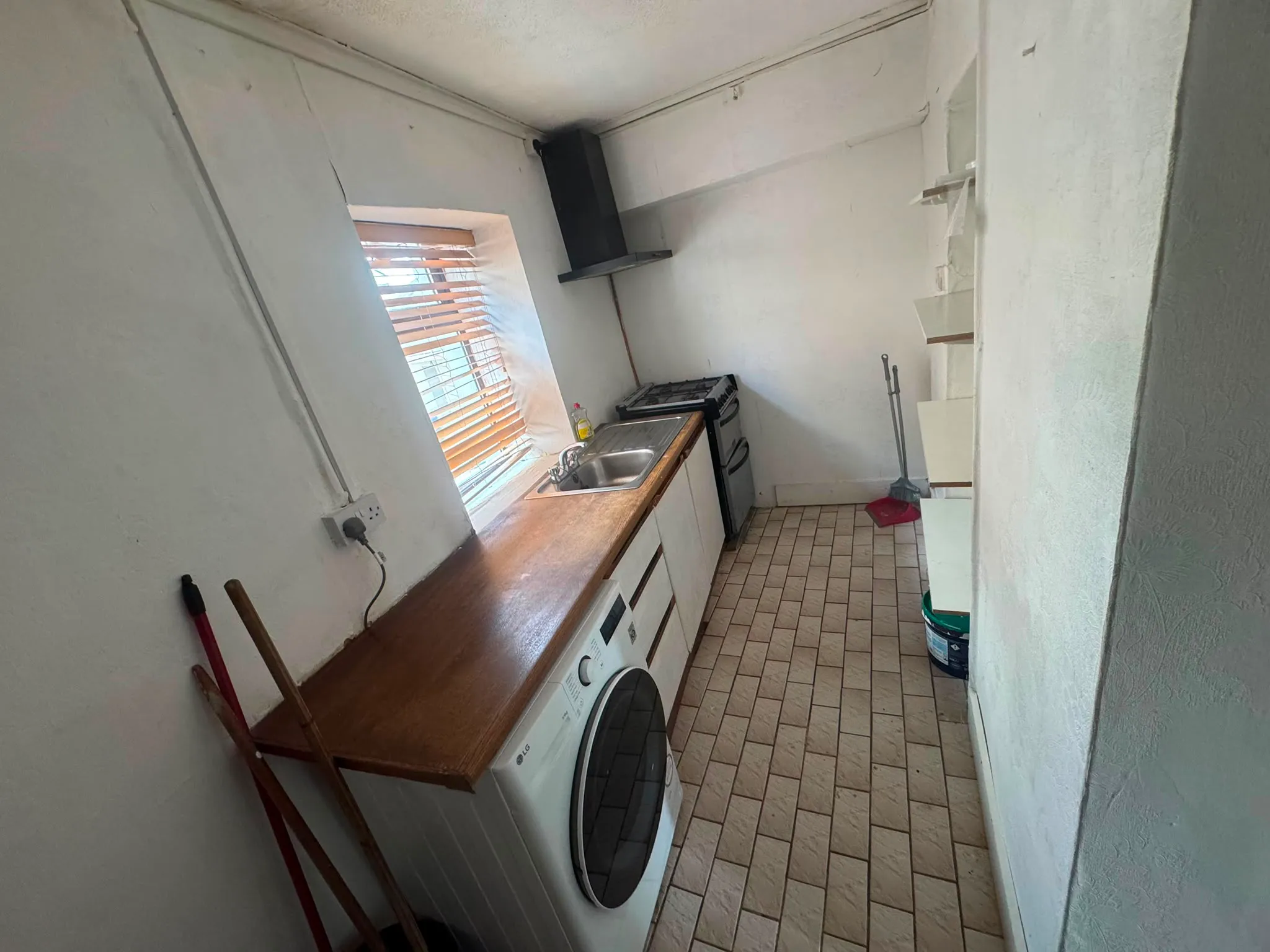 Narrow kitchen with a wooden countertop, sink, washing machine, freestanding stove, window with blinds and an extractor hood.