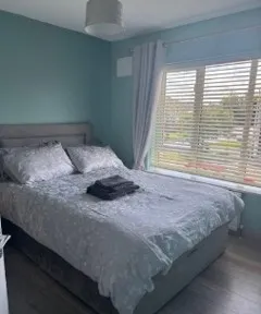 Bedroom with a double bed dressed in patterned bedding, folded towels on the bed, a large window with blinds and curtains, and a ceiling light.