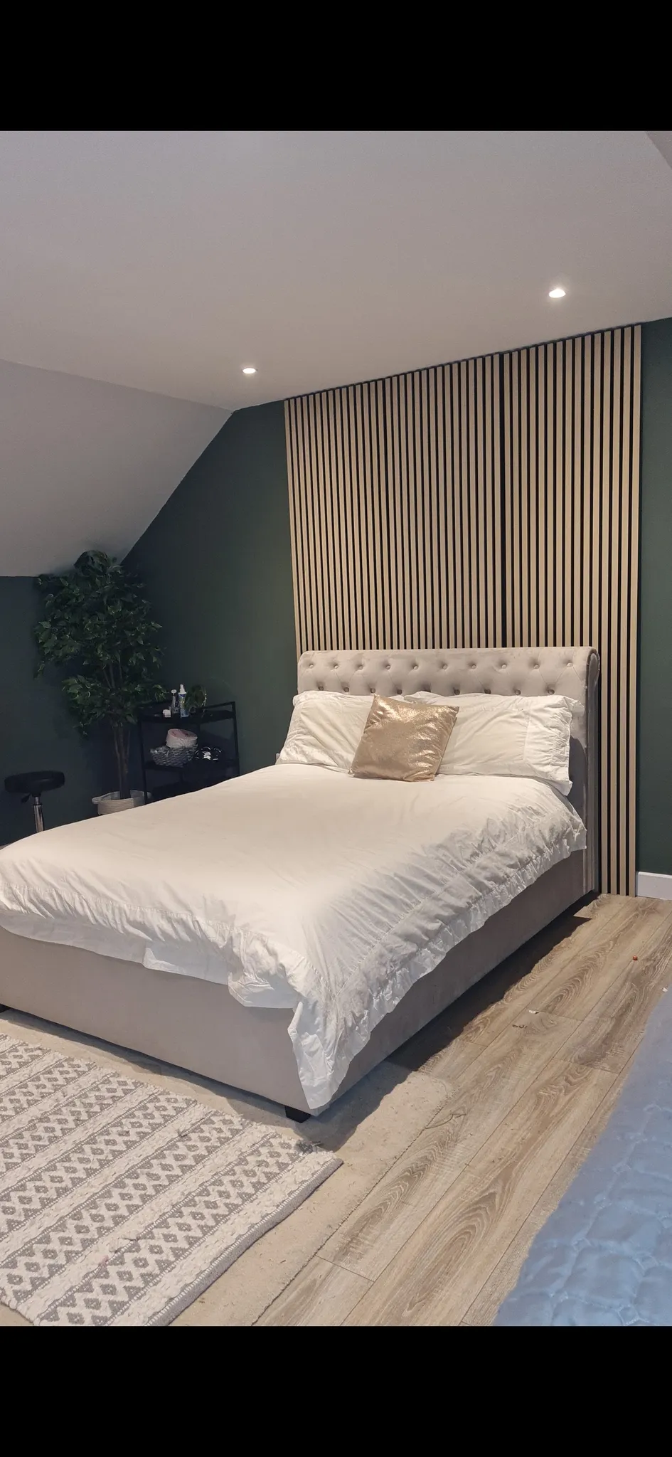 Bedroom with a queen-size bed with beige upholstered headboard and white bedding, dark teal accent wall with vertical wood slat paneling, recessed ceiling lights, wooden flooring, area rug, and potted plant on dark night