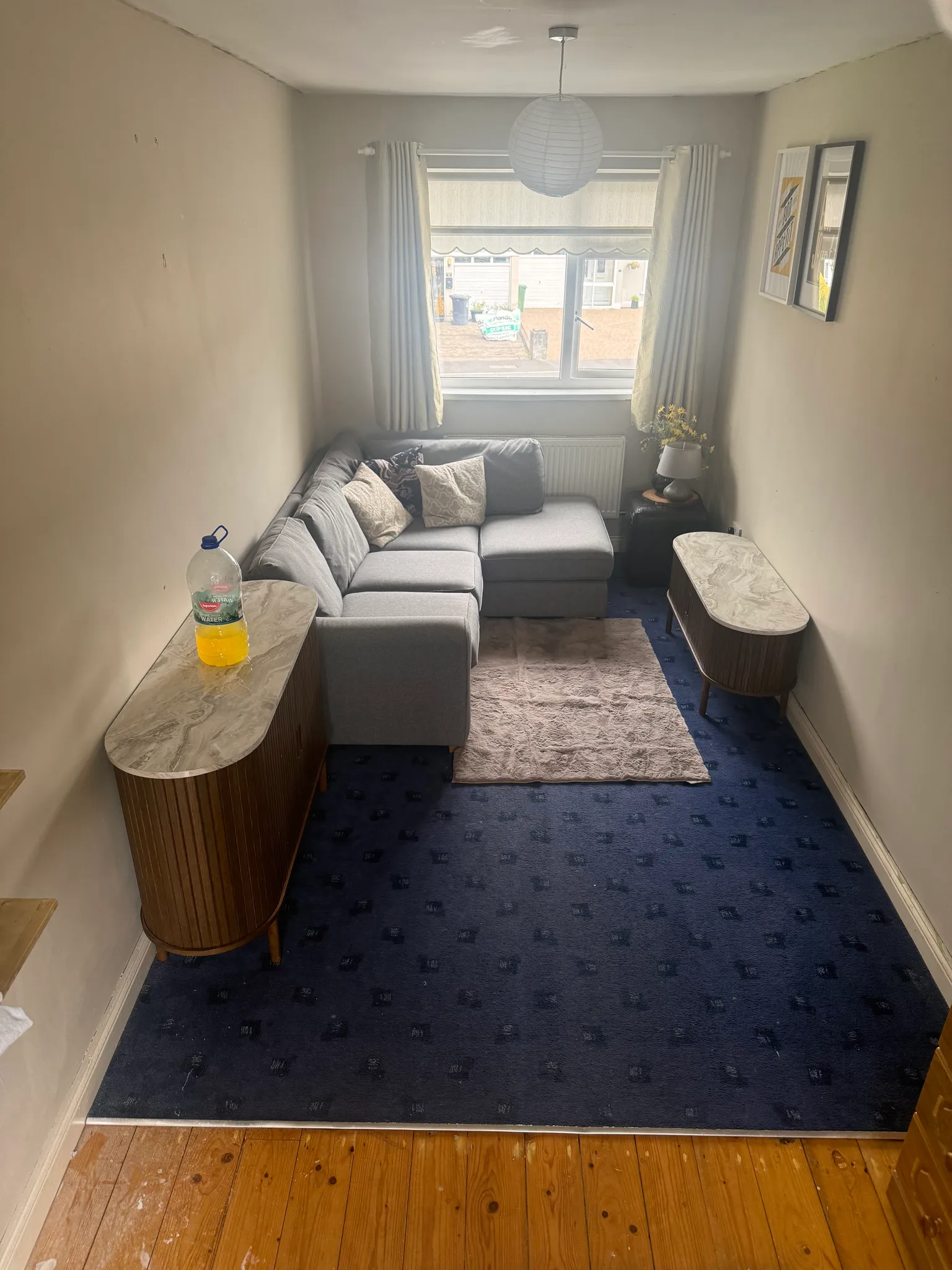 Living room with an L-shaped grey sofa under a window, small side tables with a lamp and flowers, rug on blue carpet and framed art on the wall.