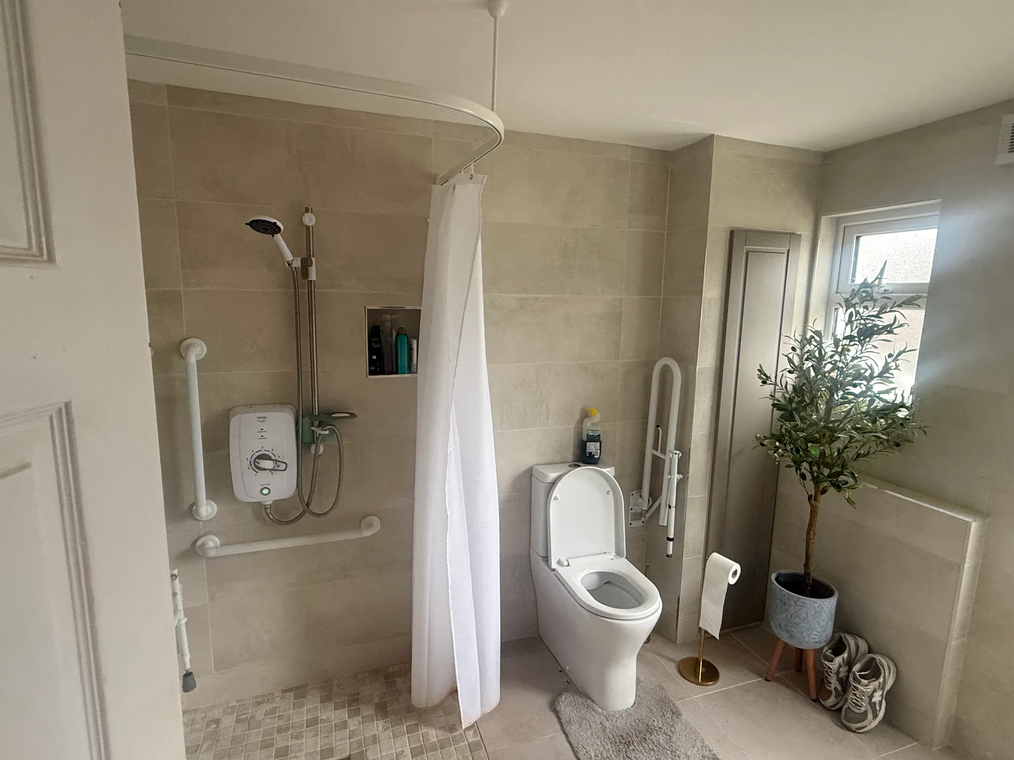 Bathroom with a wall-mounted shower and white shower curtain, a toilet with open lid, grab bars on the walls, a small window, and a potted plant.