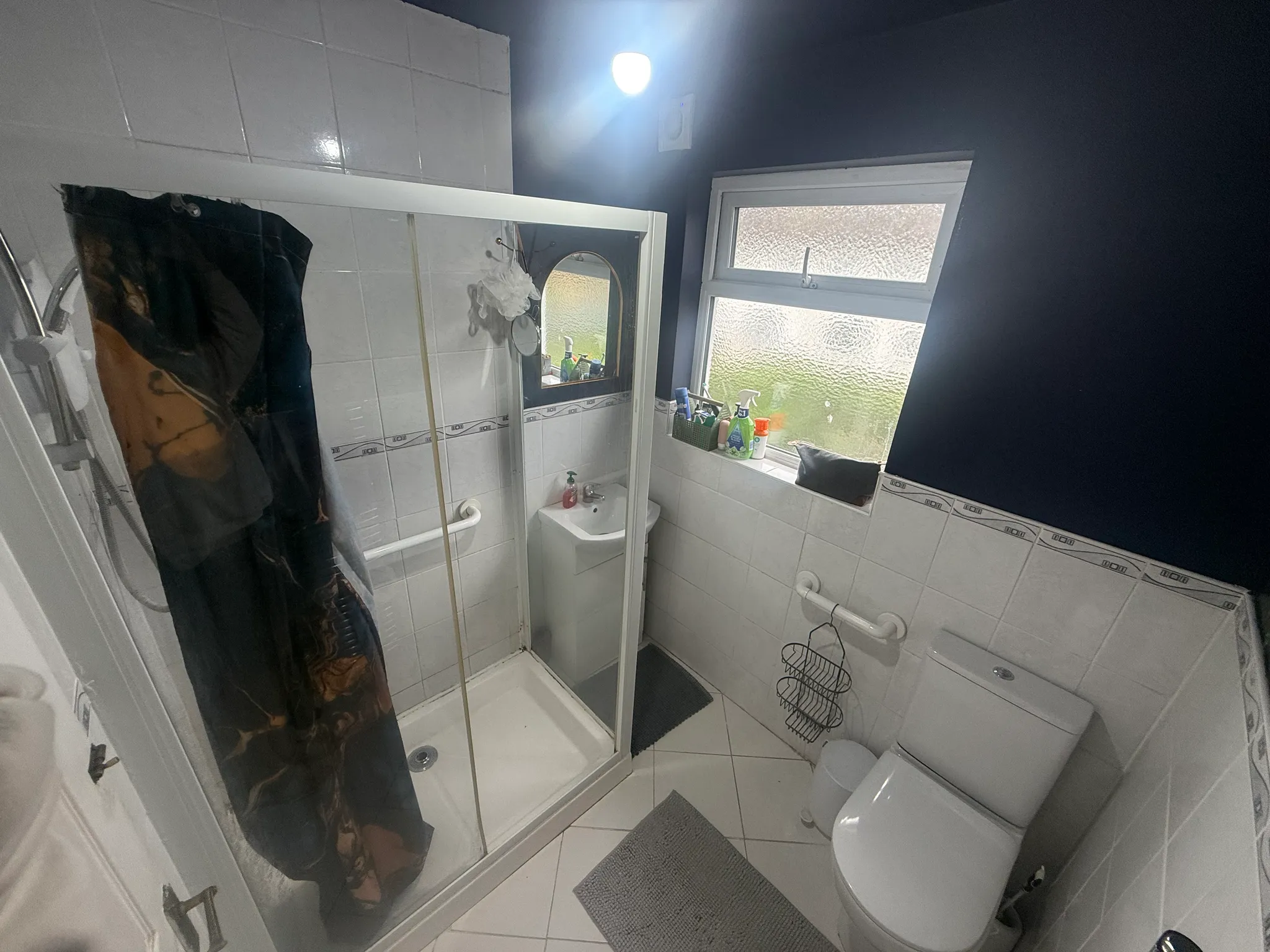 Bathroom with a corner shower enclosure, shower curtain, small sink with mirror, toilet, frosted window, and toiletries on the windowsill.