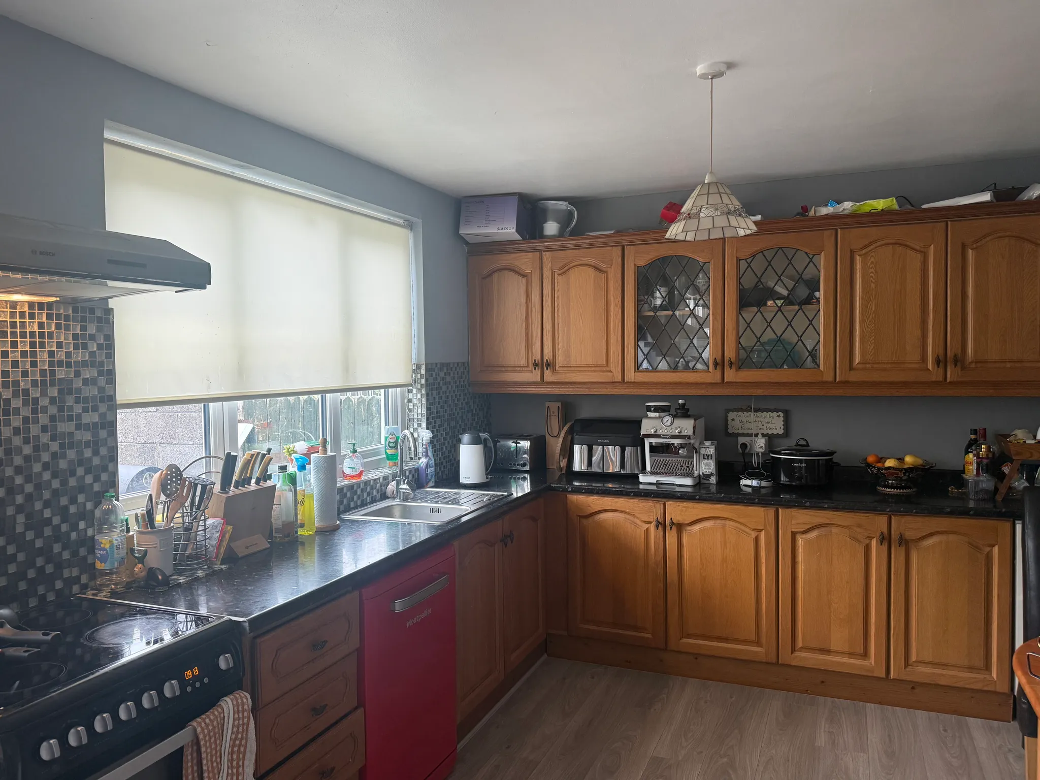Kitchen with wooden upper and lower cabinets, black countertop, sink under a window with a closed roller blind, stove and extractor hood, and appliances on the counter.