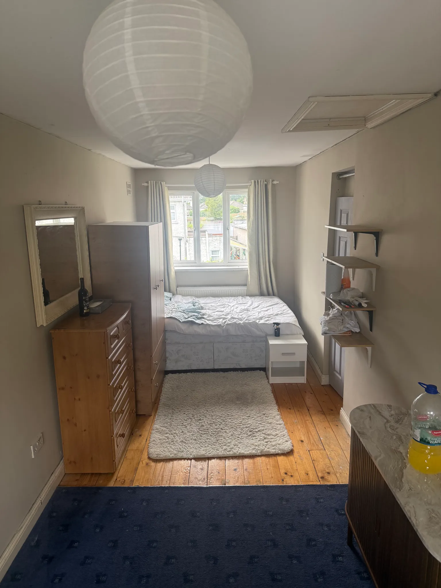Bedroom with a double bed under a window, wardrobe and chest of drawers, wall shelves and two hanging paper lantern lights