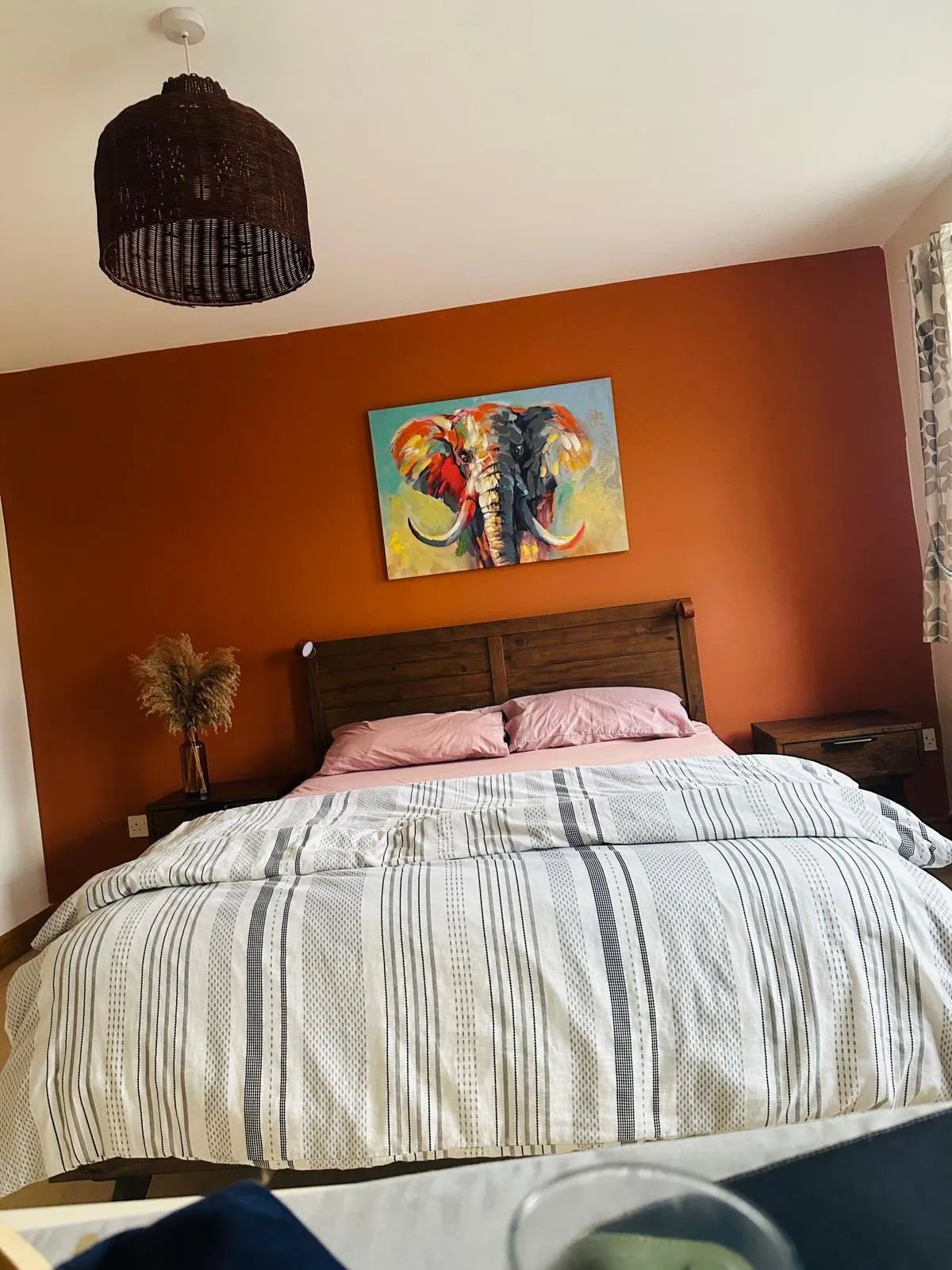 Bedroom with a double bed, wooden headboard, striped duvet, bedside tables, and a colorful elephant painting on an orange accent wall.