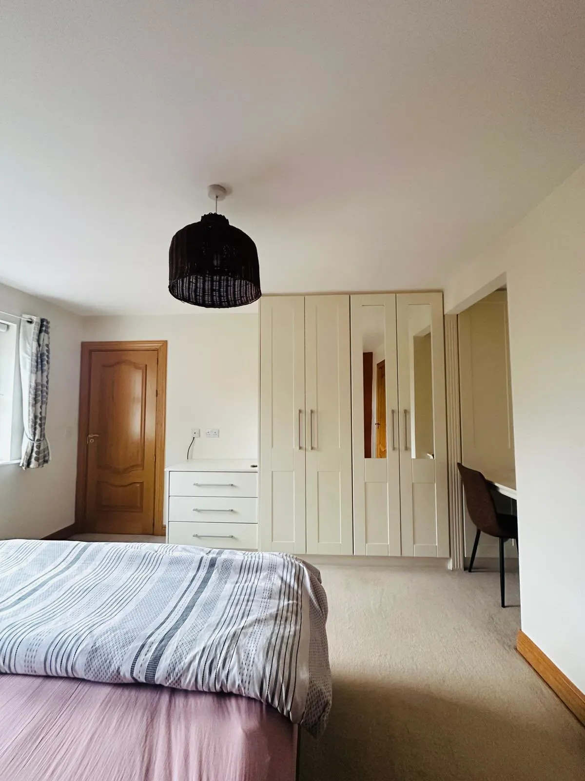 Bedroom with a bed in the foreground, built-in wardrobe with mirrored panel, chest of drawers, window with curtains, wooden door and a desk with chair in a alcove.