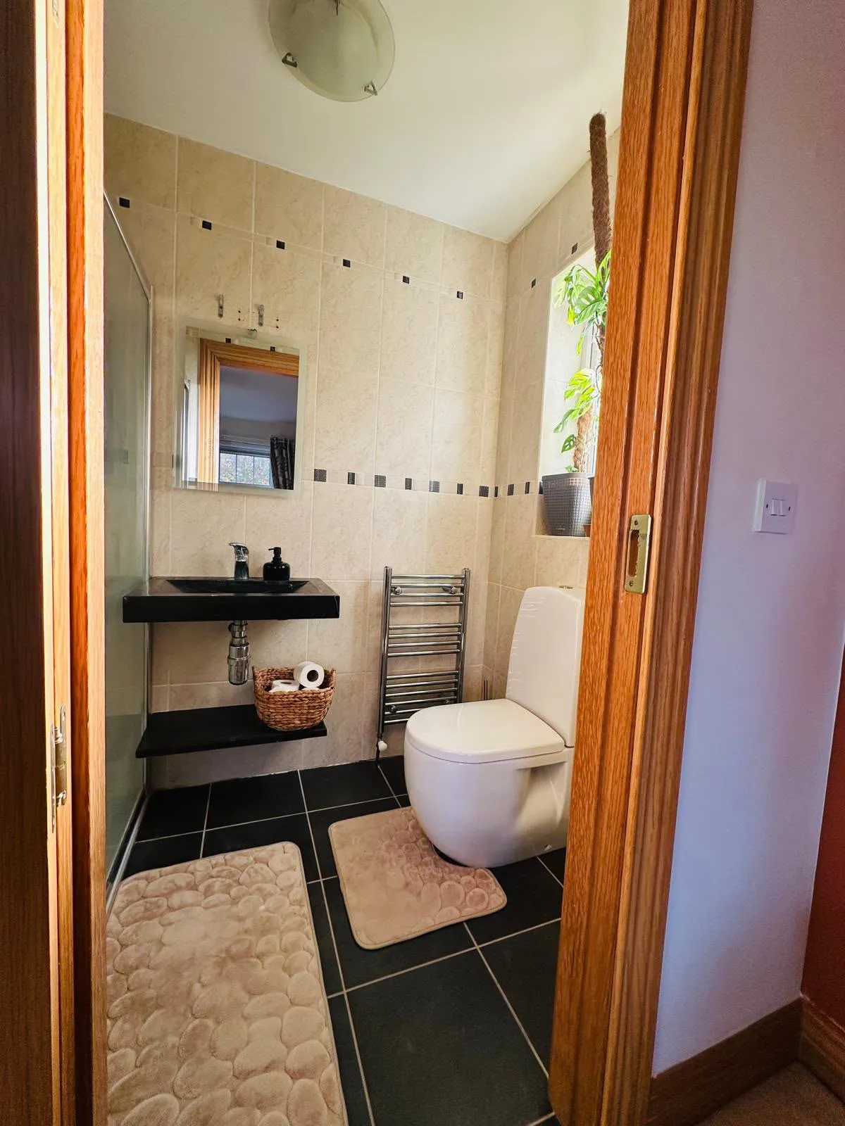Small bathroom with a toilet, wall-mounted black sink and mirror, heated towel rail, window with a potted plant, and beige tiled walls