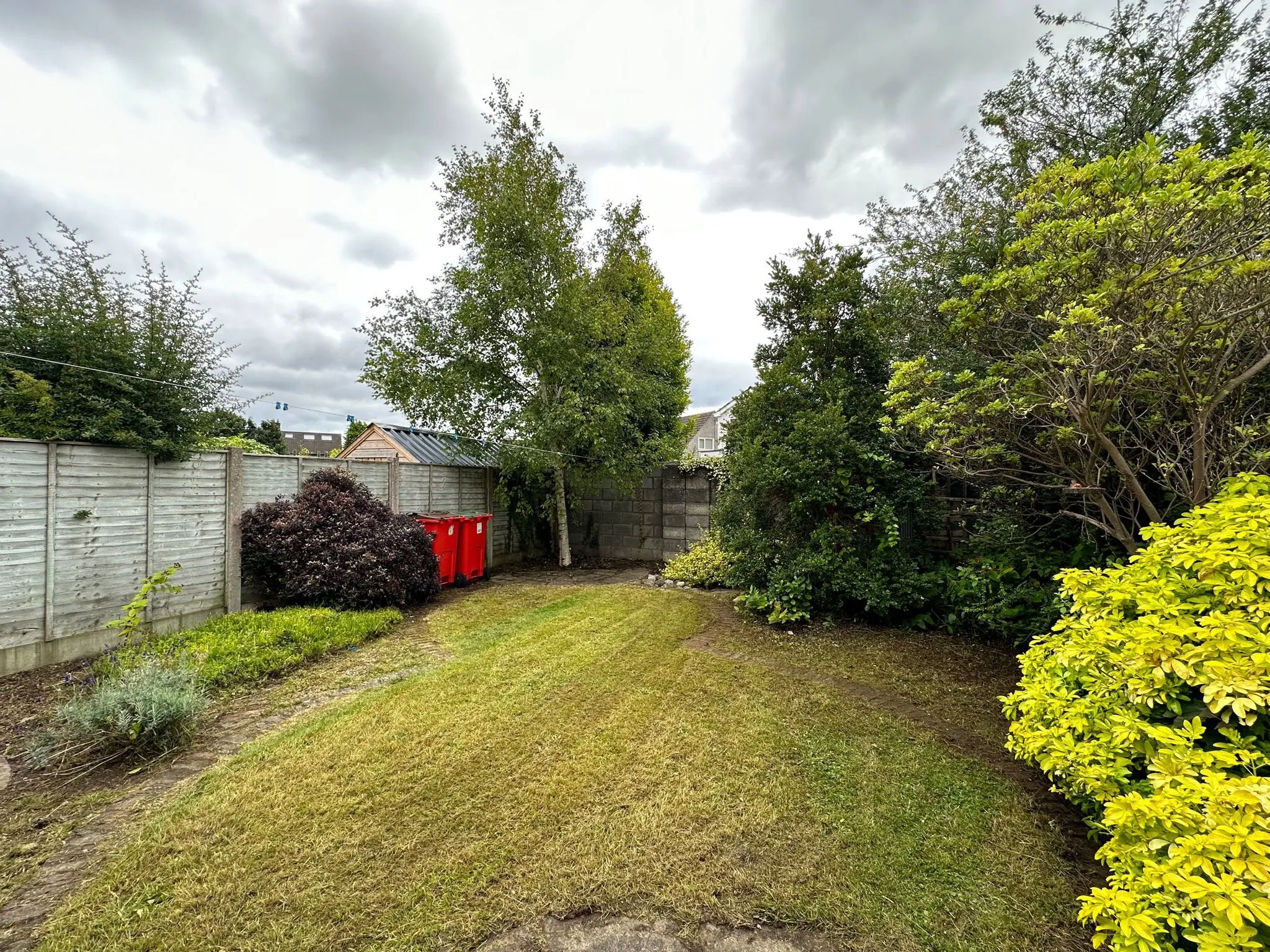 Backyard garden with a lawn, wooden fence, trees and shrubs, two red waste bins near a corner and an overcast sky.