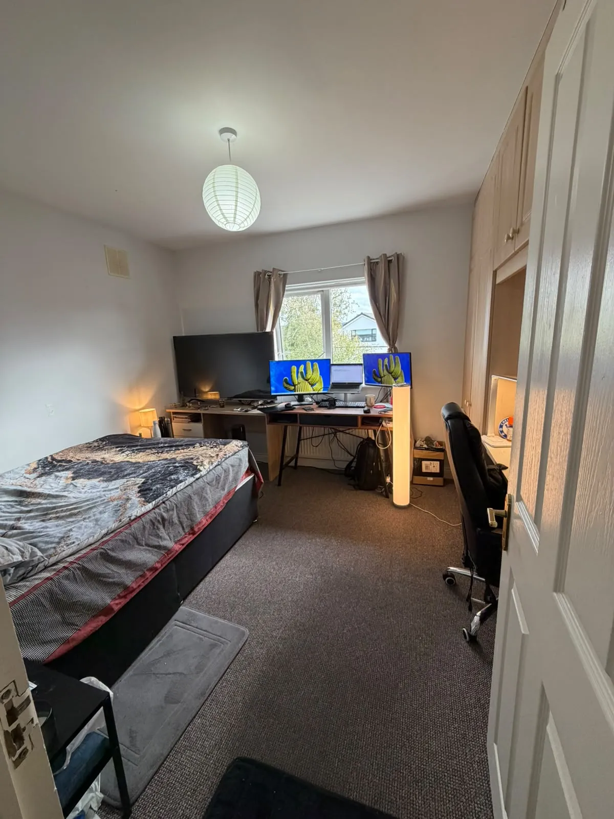 Bedroom with a bed on the left, a desk with three monitors under a window, an office chair, built-in wardrobes on the right and a ceiling light.