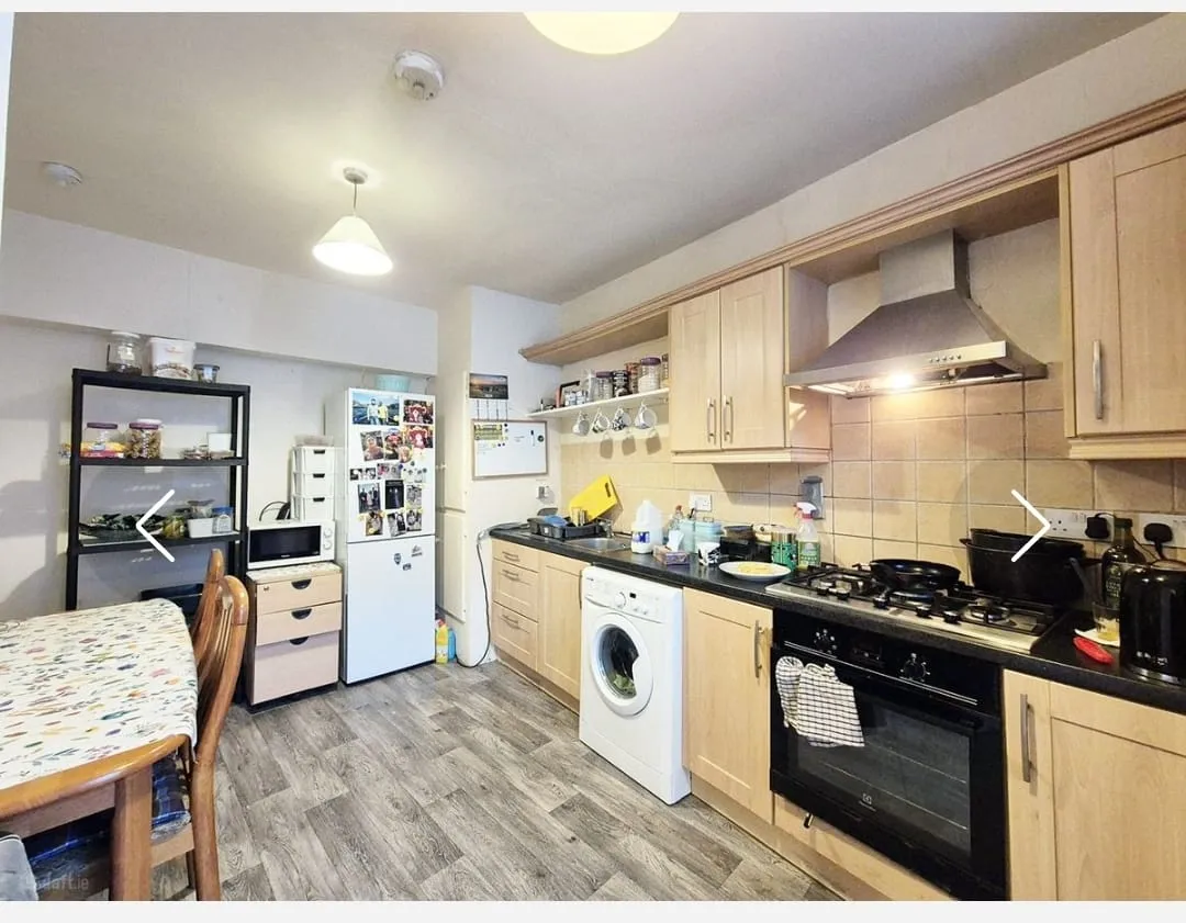 Kitchen with wooden cabinets, gas stove and oven with extractor hood, countertop with sink and small appliances, a washing machine under the counter, a refrigerator with photos on it, shelving unit and a dining table at