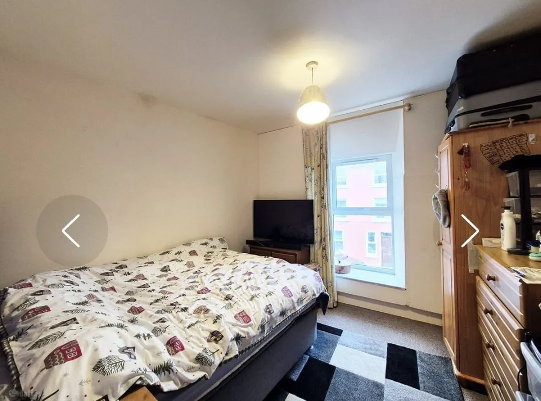Bedroom with a double bed, patterned duvet, window with curtain, freestanding TV on a low stand, tall wardrobe and chest of drawers.