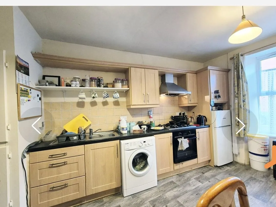 Kitchen with light wood cabinets, sink, washing machine under the counter, gas oven with range hood, refrigerator, countertop items and a window with curtain.