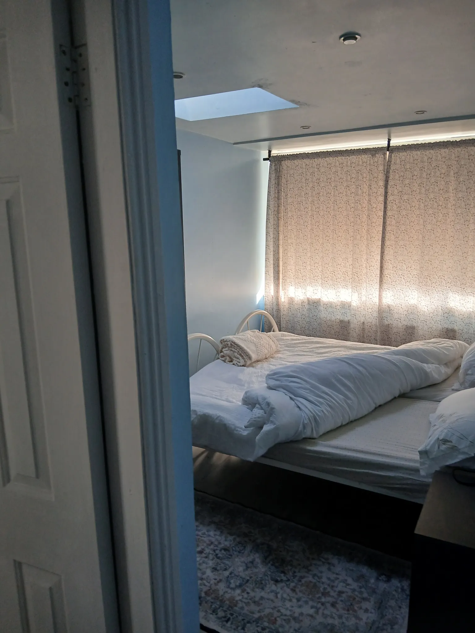Bedroom with an unmade double bed, patterned curtains covering a window, and a ceiling skylight visible.