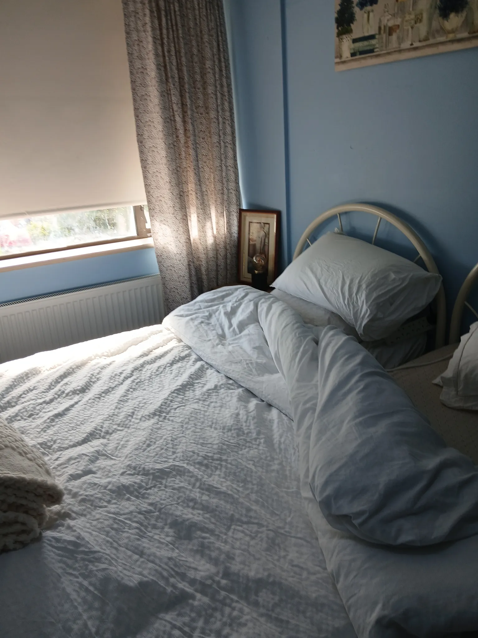 Bedroom corner with a single metal headboard bed, white pillows and duvet, a window with a blind and curtain, radiator and a framed picture against a blue wall.