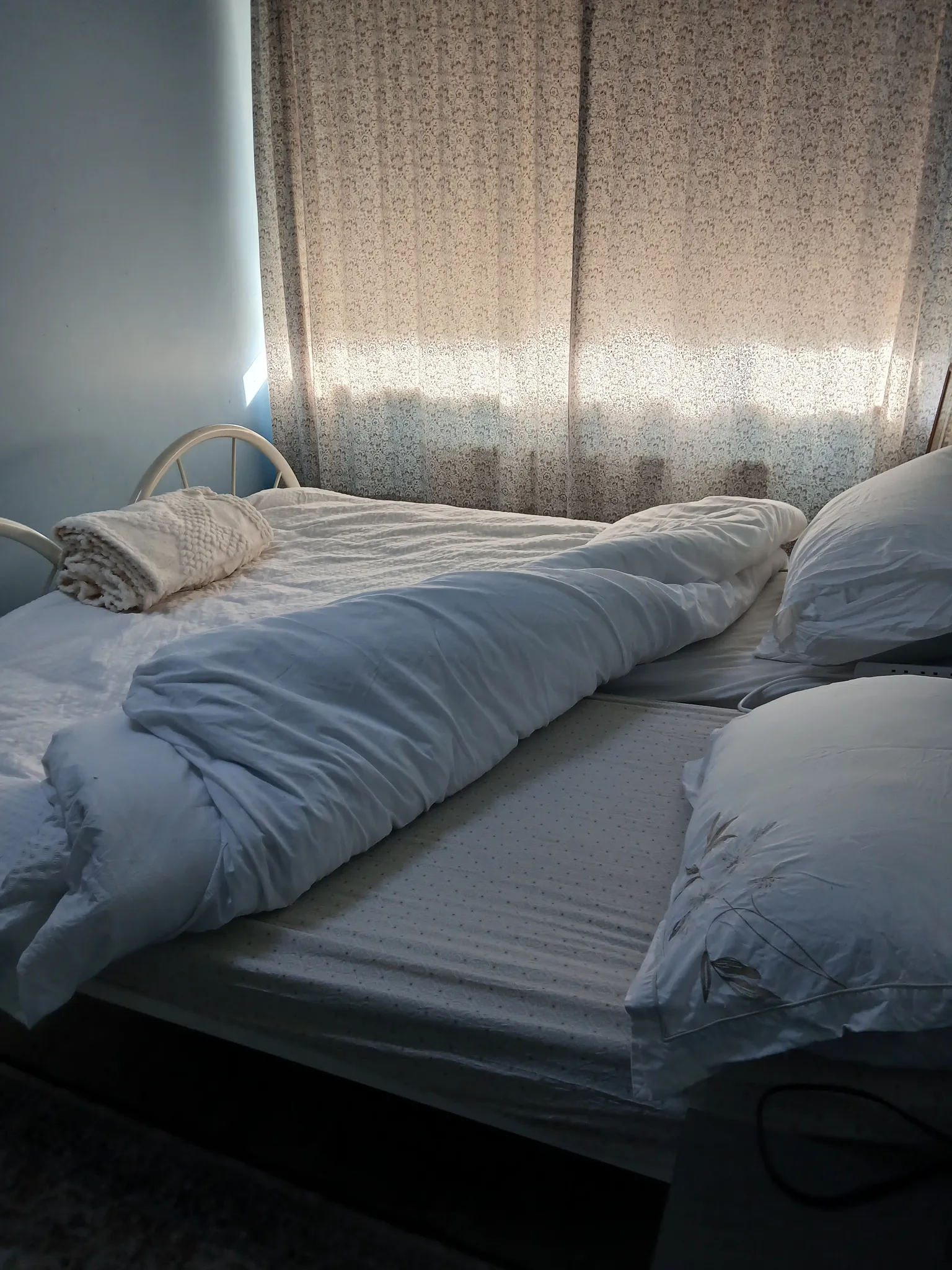 Bedroom with a double bed, white bedding and pillows, folded blanket on the foot of the bed, and patterned curtains covering a window.