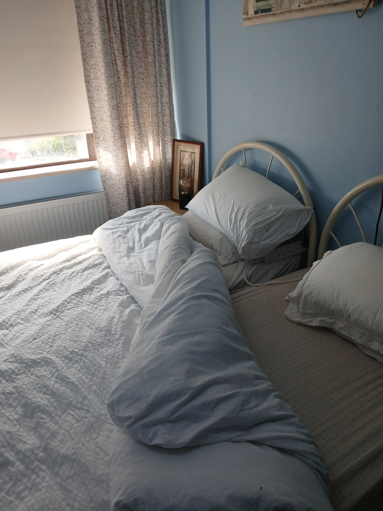 Bedroom with two single metal-framed beds, white bedding and pillows, a window with a blind and patterned curtain, and a small bedside table holding a lamp and framed picture.