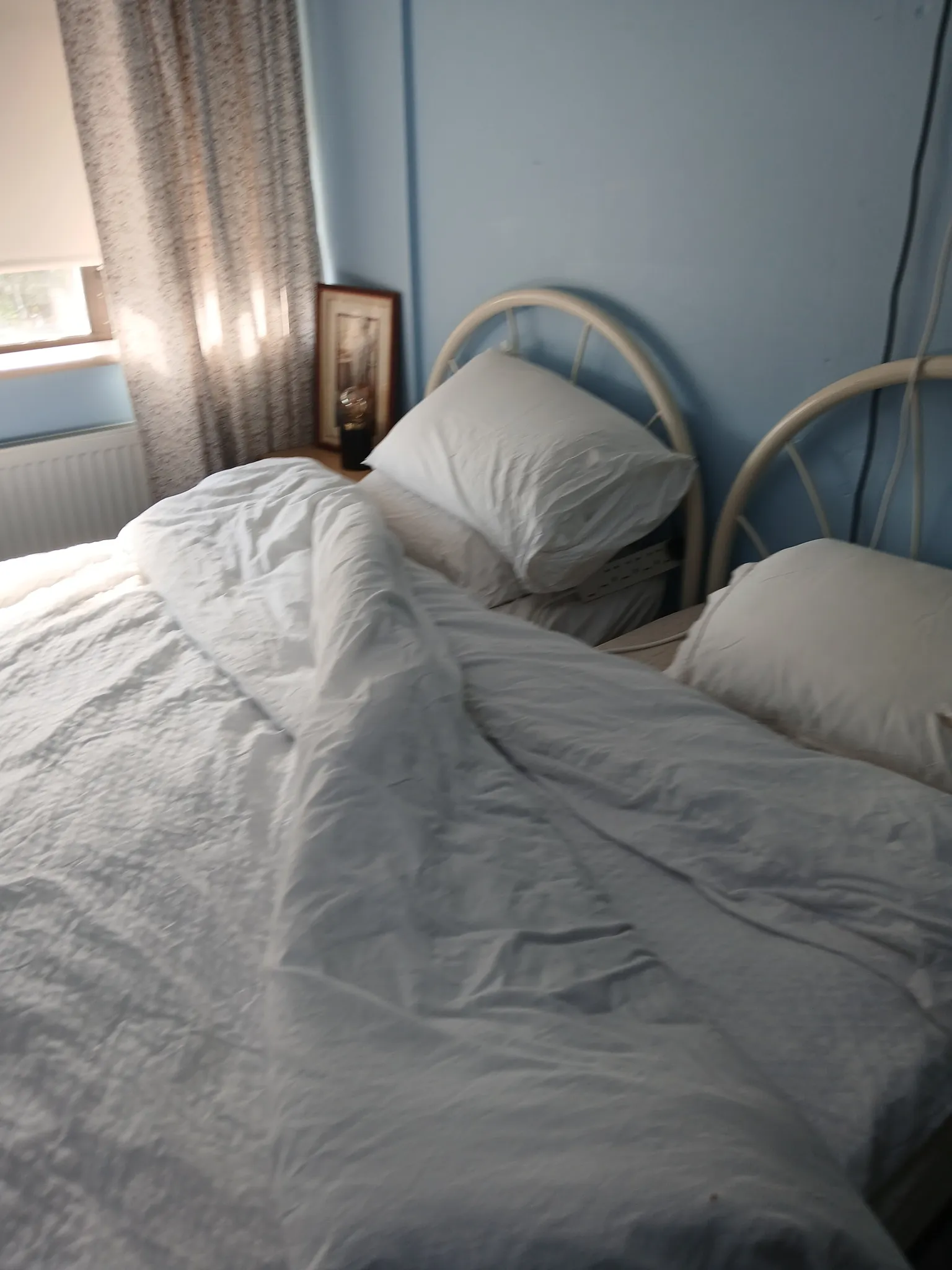 Bedroom with two single metal headboards, white bedding and pillows, blue wall, window with curtain and a small bedside table with framed picture and lamp.