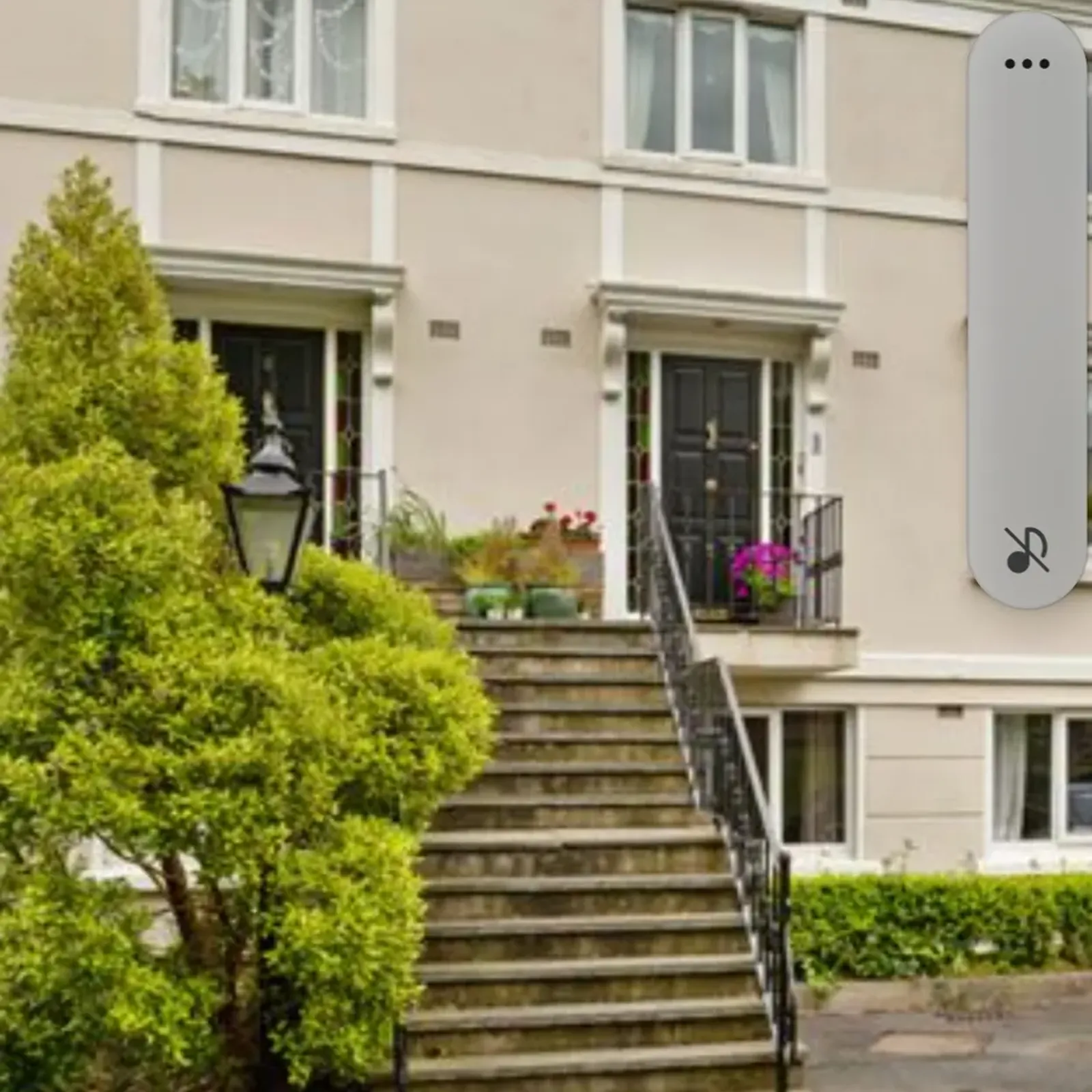 Exterior view of a two-story building with a central stone staircase leading to two black front doors, windows above, metal railings, potted plants and a lamp post to the left.