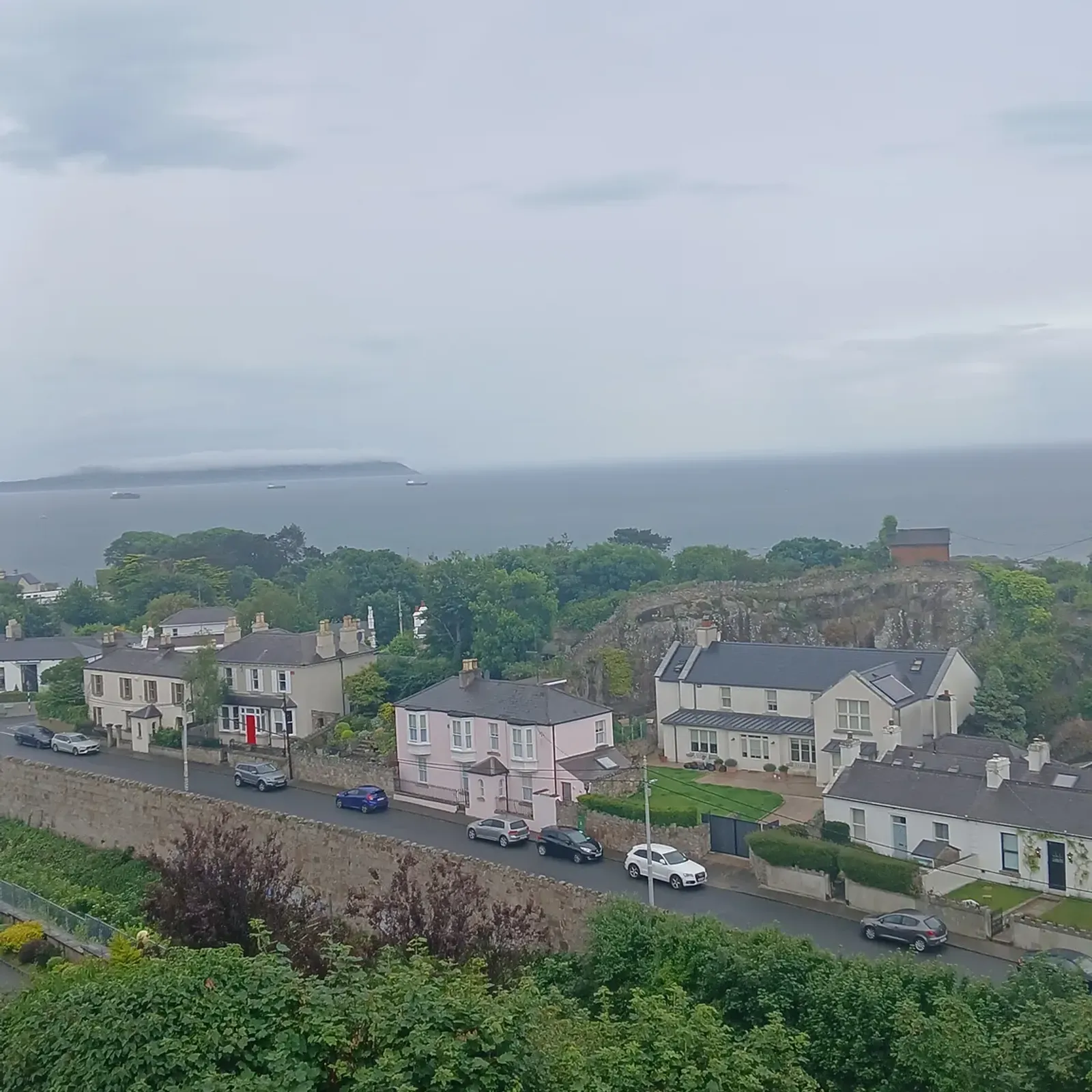 Exterior view from a higher level showing residential houses and a street with parked cars, green shrubs in the foreground and the sea with a distant shoreline under a cloudy sky.