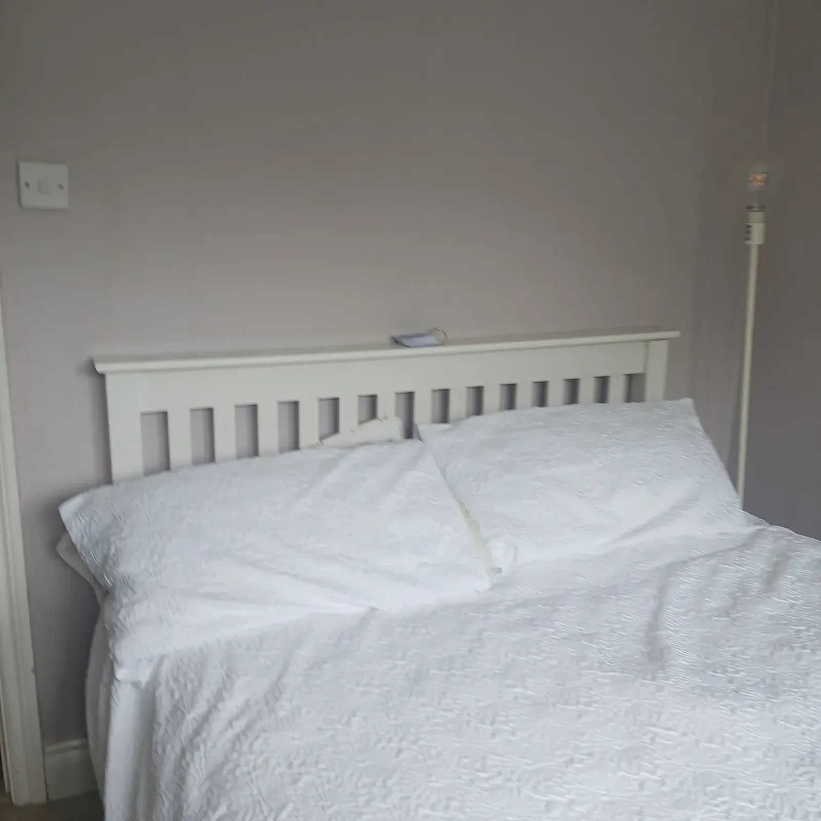 Bedroom with white bed and pillows, white slatted headboard, floor lamp to the right, and wall switch to the left.