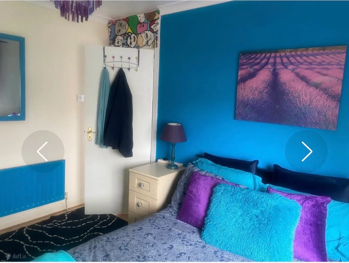 Bedroom with a double bed against a bright blue accent wall, bedside table with lamp, colorful pillows, wall art, and a door with hanging coats.