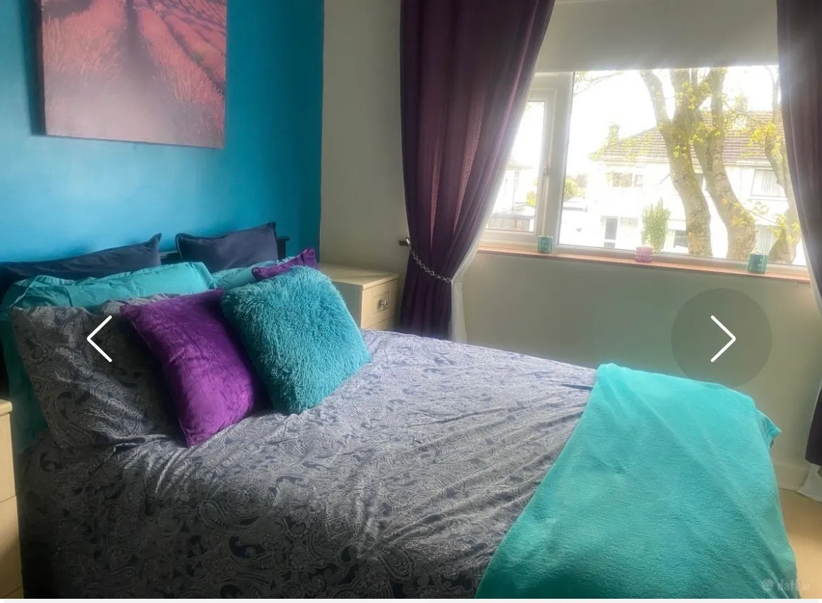 Bedroom with a double bed, teal and purple throw pillows, teal blanket, bedside table, window with dark curtains, and a teal-painted accent wall with wall art.