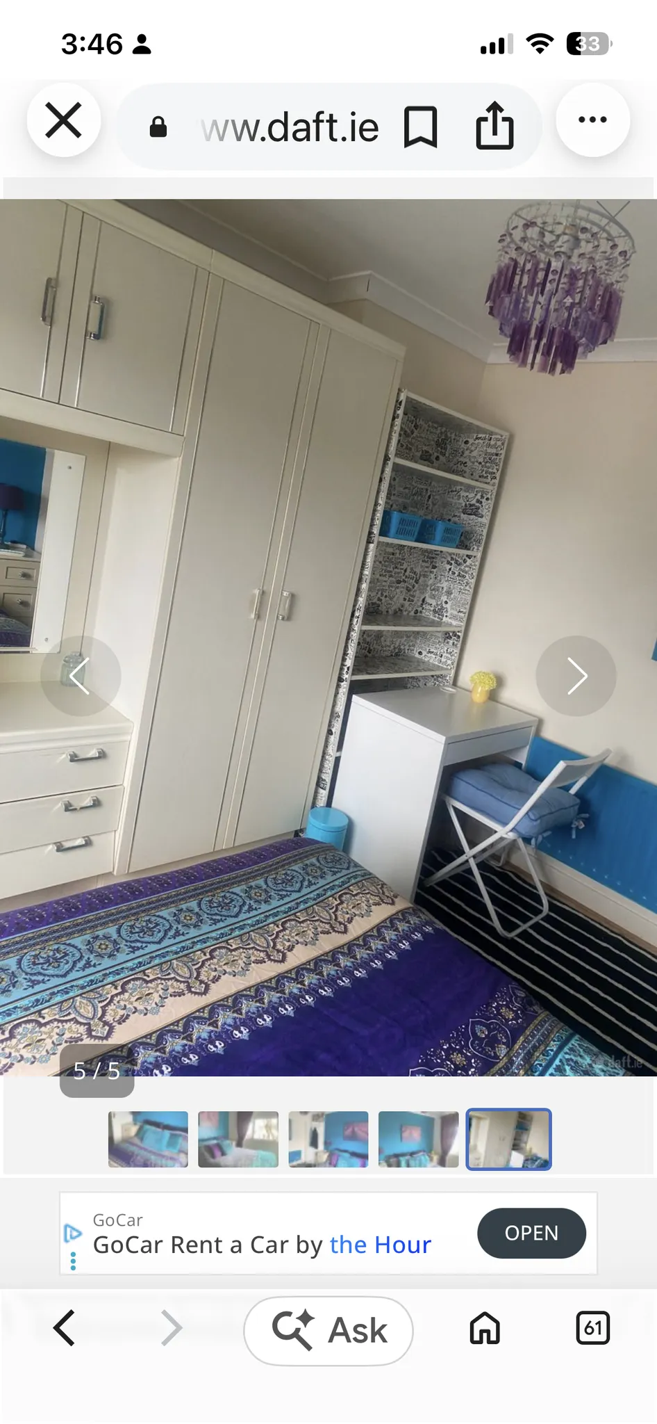 Bedroom corner with a double bed (purple patterned bedding), built-in wardrobe with mirror, small white desk and folding chair, open shelving, and a purple hanging light fixture.