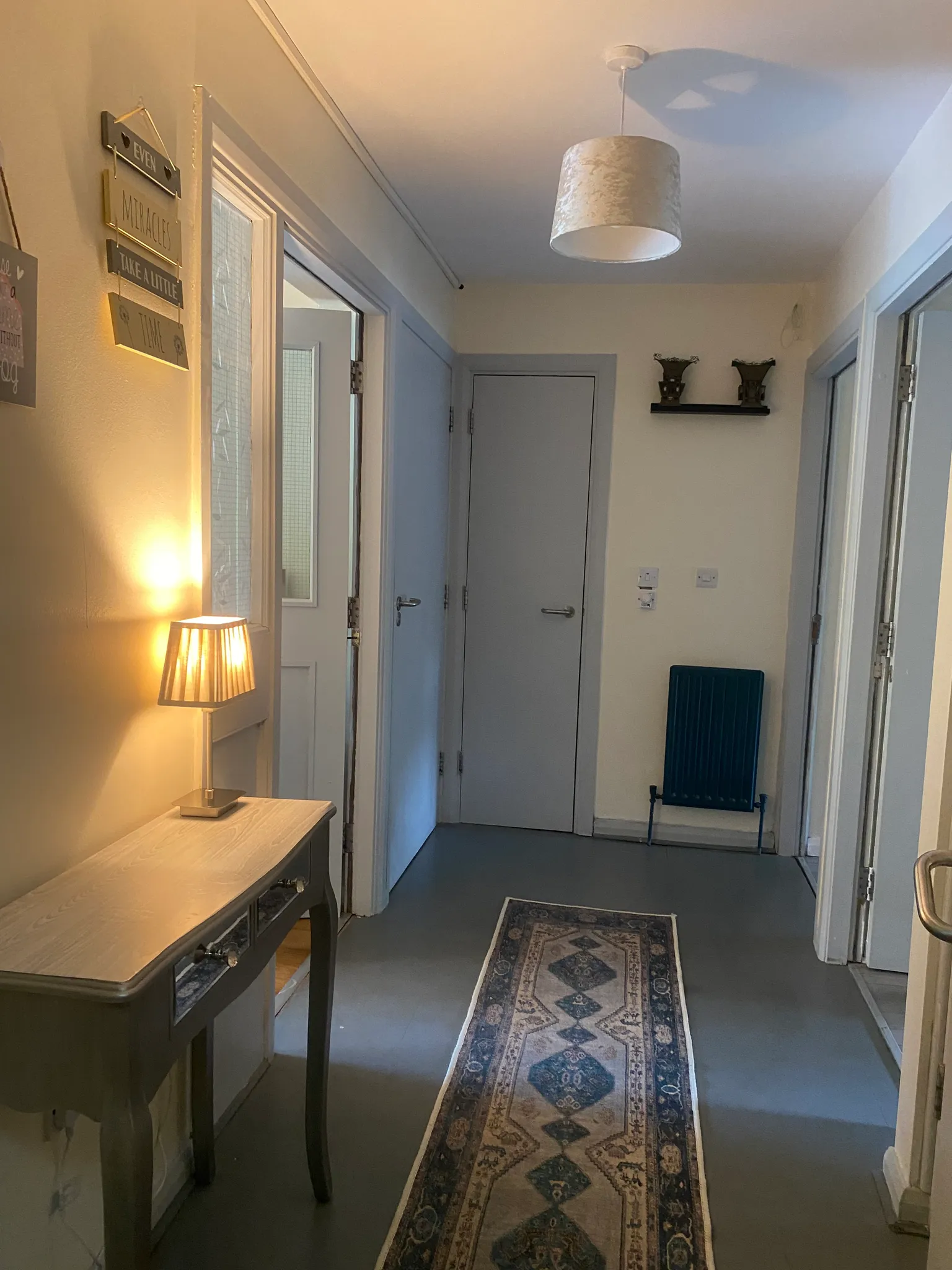 Hallway with a narrow patterned runner rug, console table and lamp on the left, multiple doors and a pendant ceiling light.