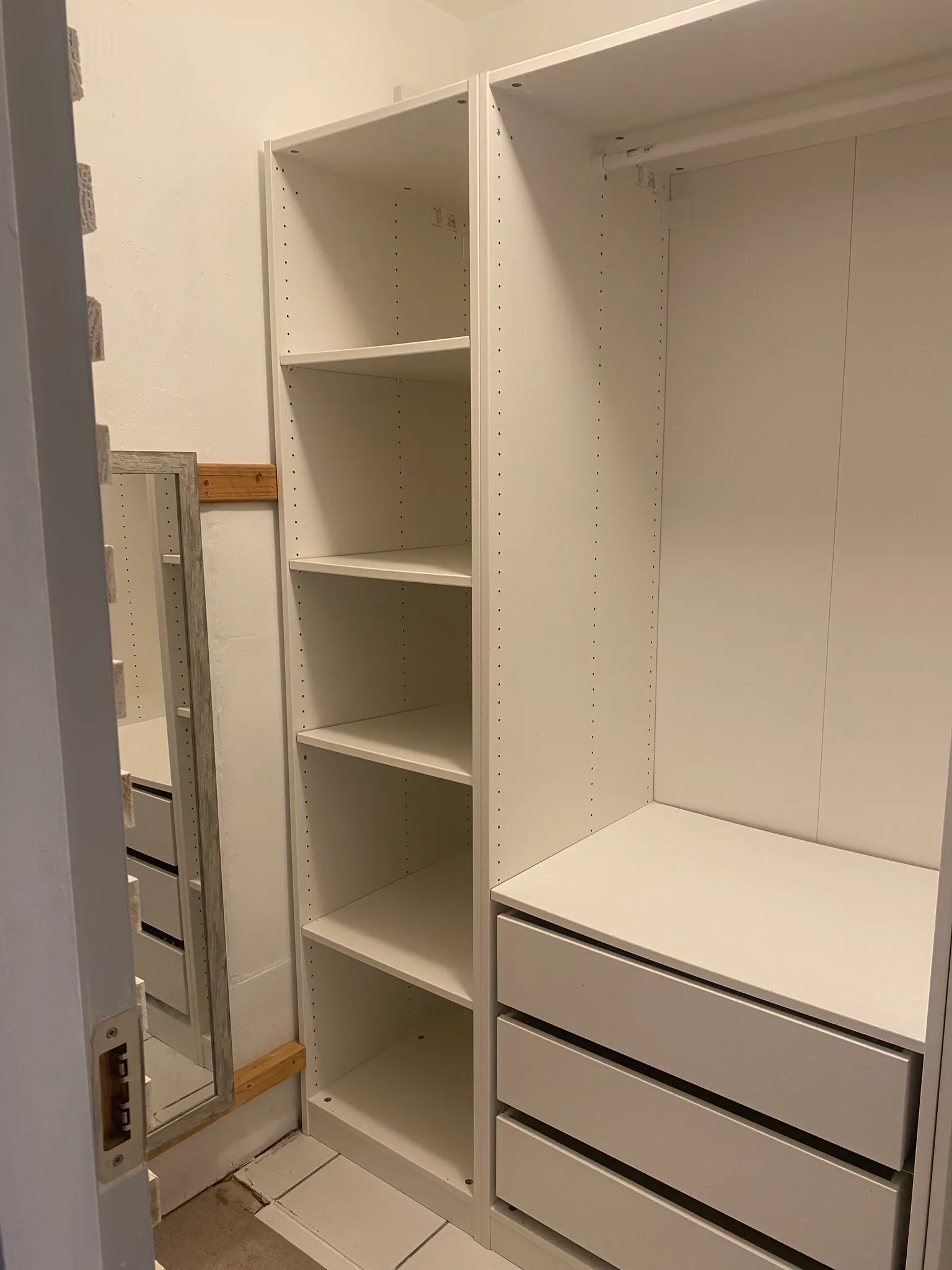 Closet area with built-in white shelves, drawers and a hanging rod, plus a leaning mirror on the left.