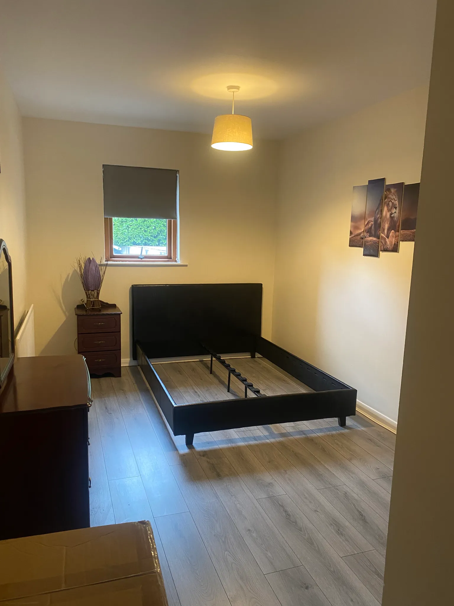 Bedroom with an empty black bed frame, small window with a closed blind, bedside table and pendant ceiling light.