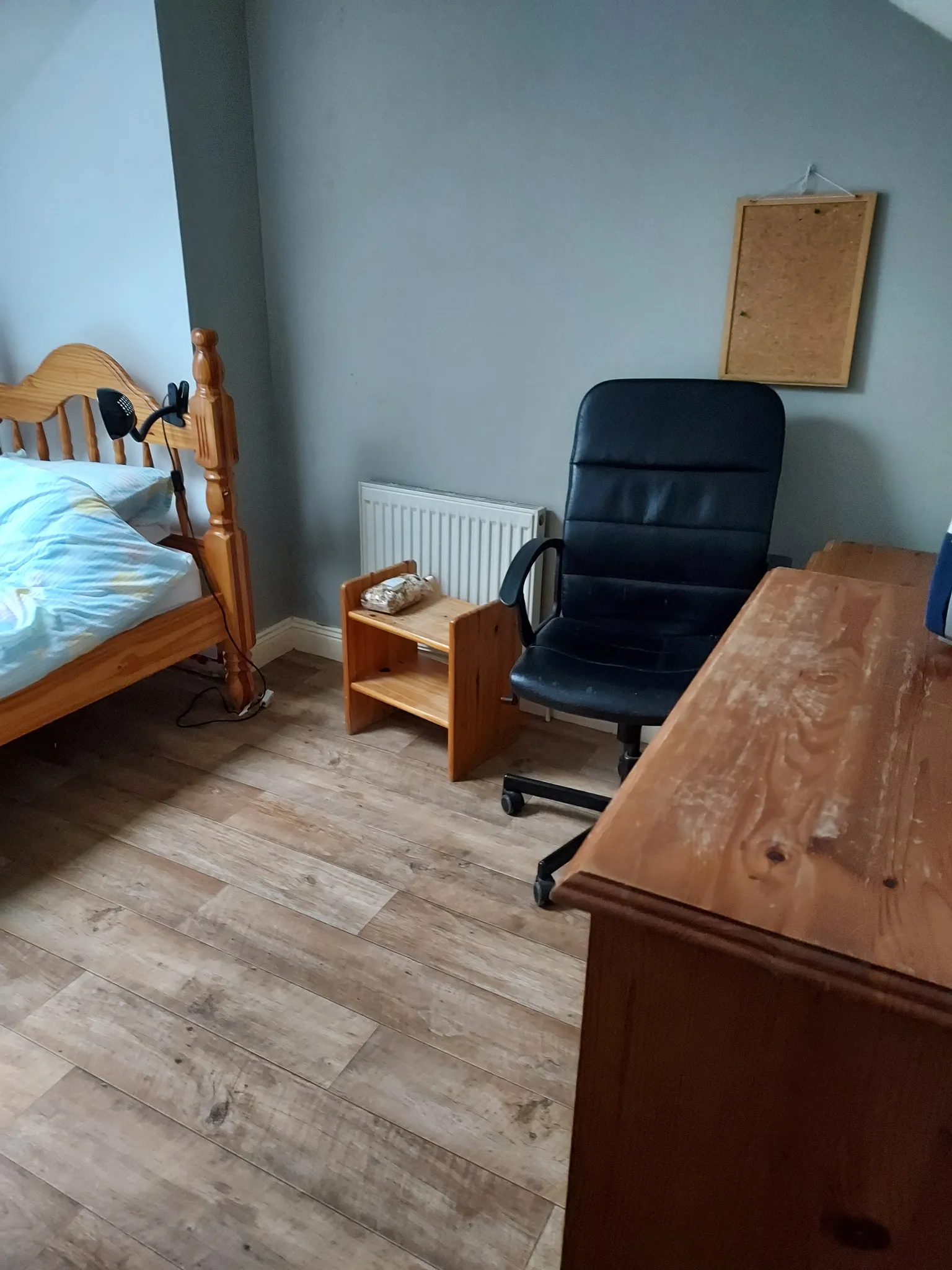 Bedroom corner with a wooden single bed, bedside lamp, small wooden shelf, black office chair, wooden desk and a cork noticeboard on a grey wall.