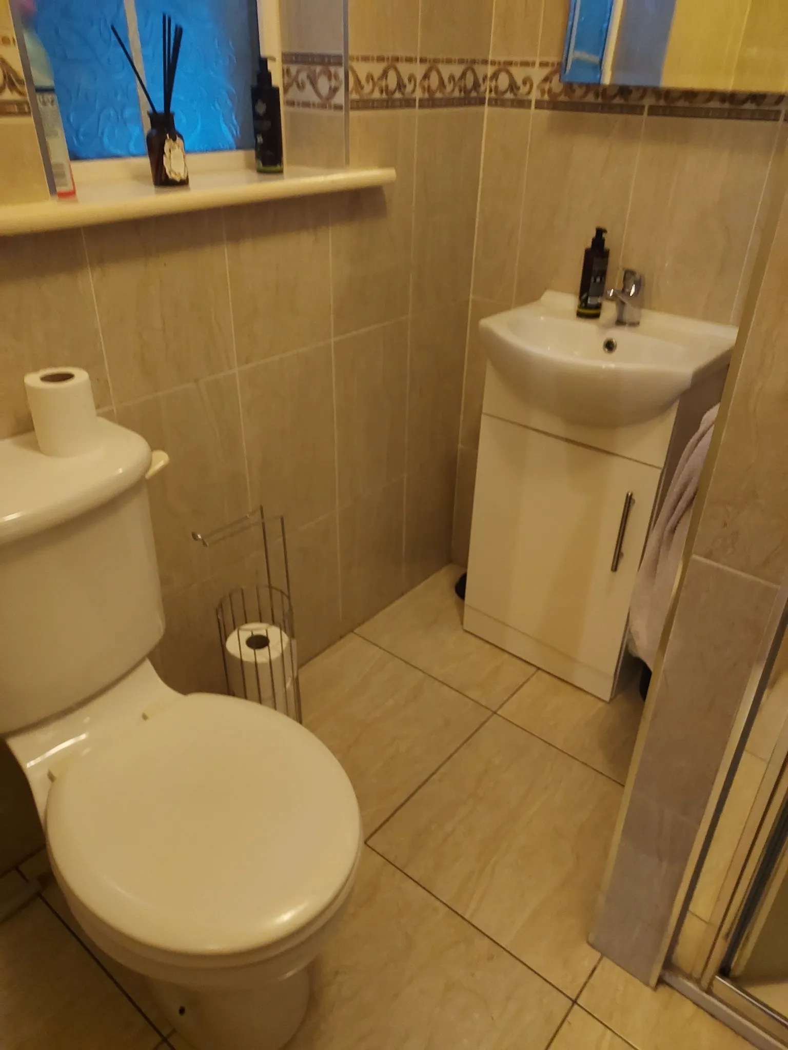 Small bathroom with a toilet, a sink cabinet, tiled walls and floor, a mirror and a window shelf with toiletries.
