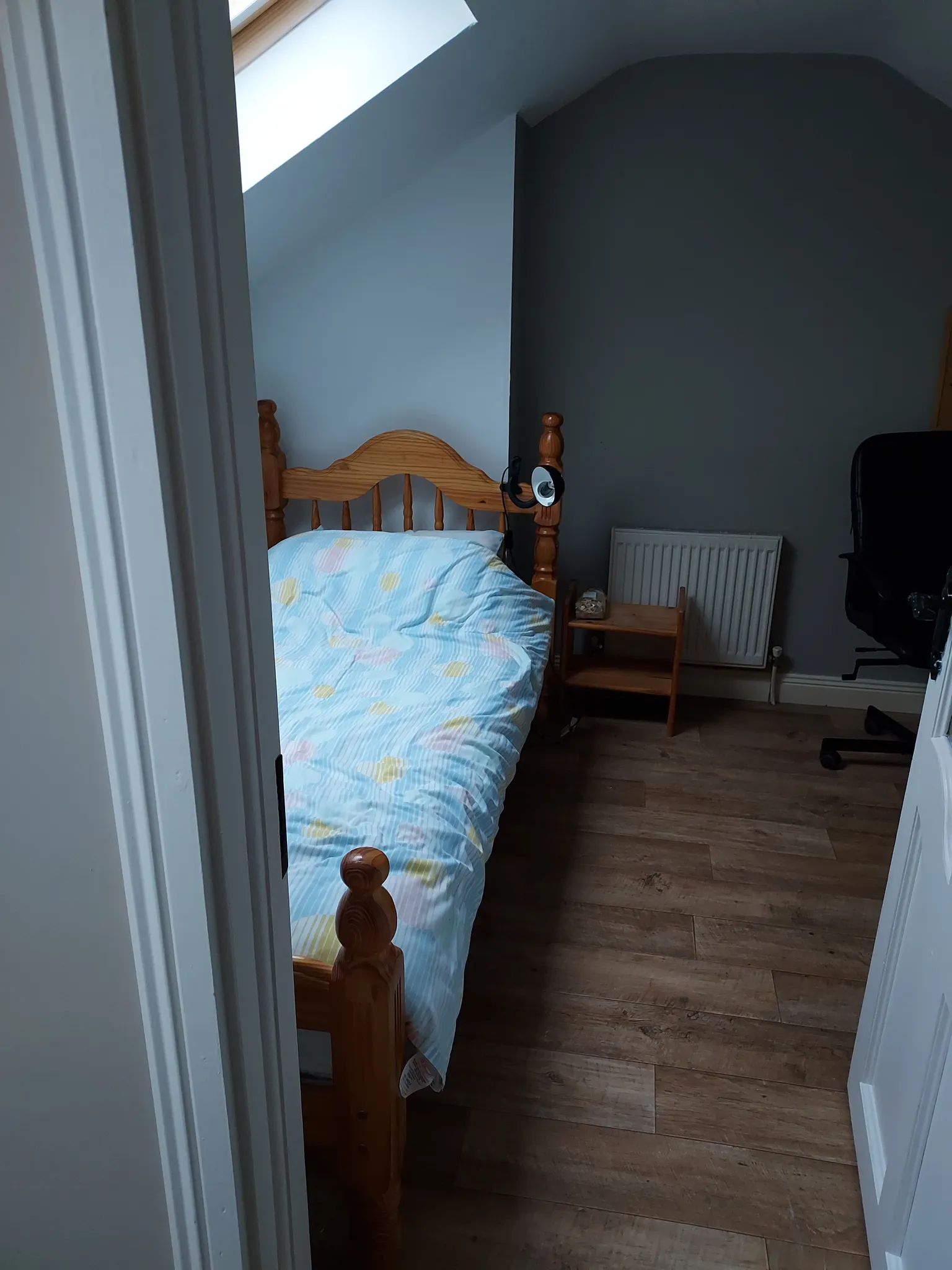 Bedroom with a single wooden bed under a sloped ceiling skylight, small bedside table, radiator and office chair partially visible