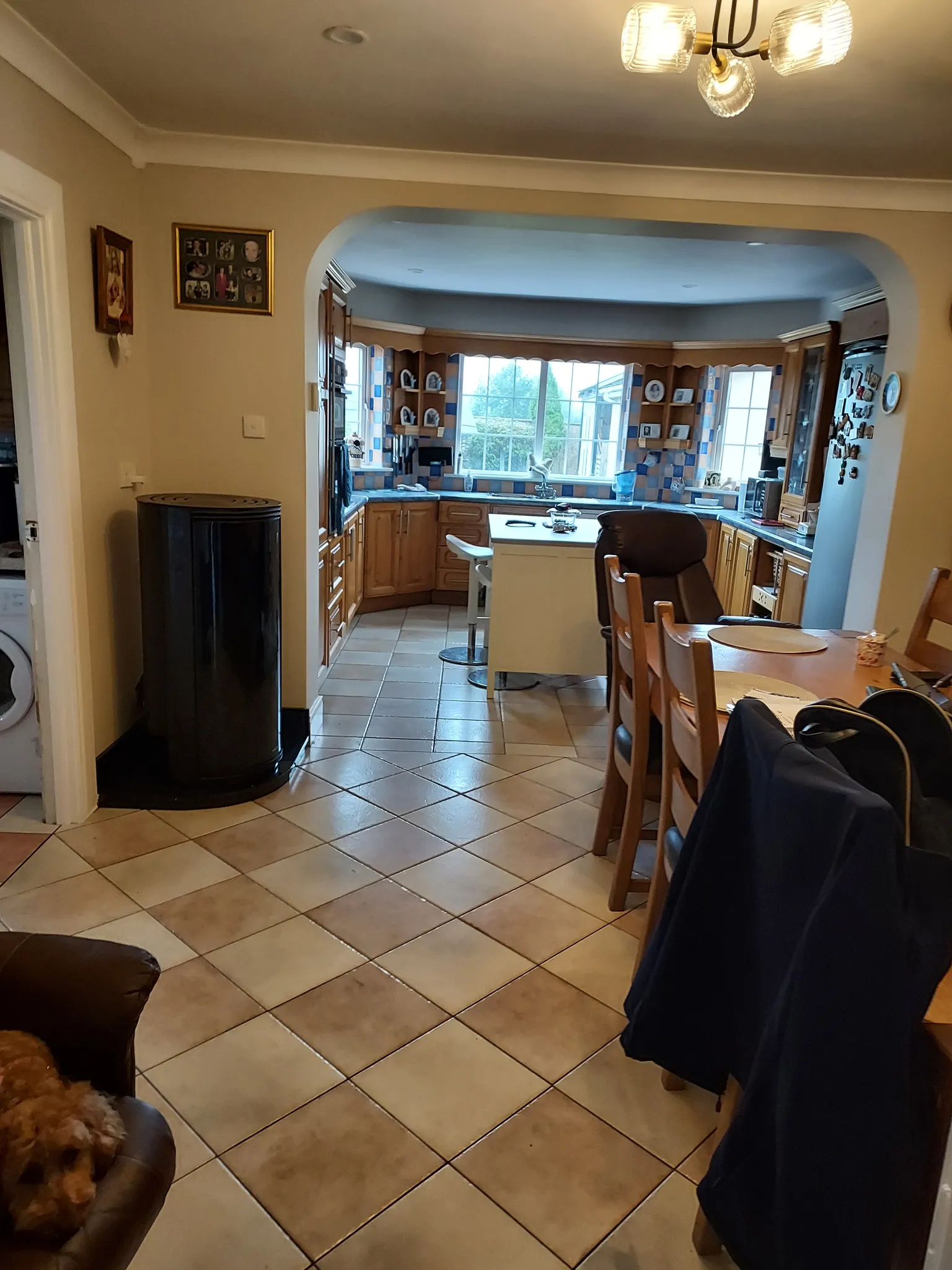 Open-plan view of a kitchen and dining area seen through an archway, with a tiled floor, wooden cabinets, central island with a stool, large window above the sink, fridge at right and dining chairs at a table
