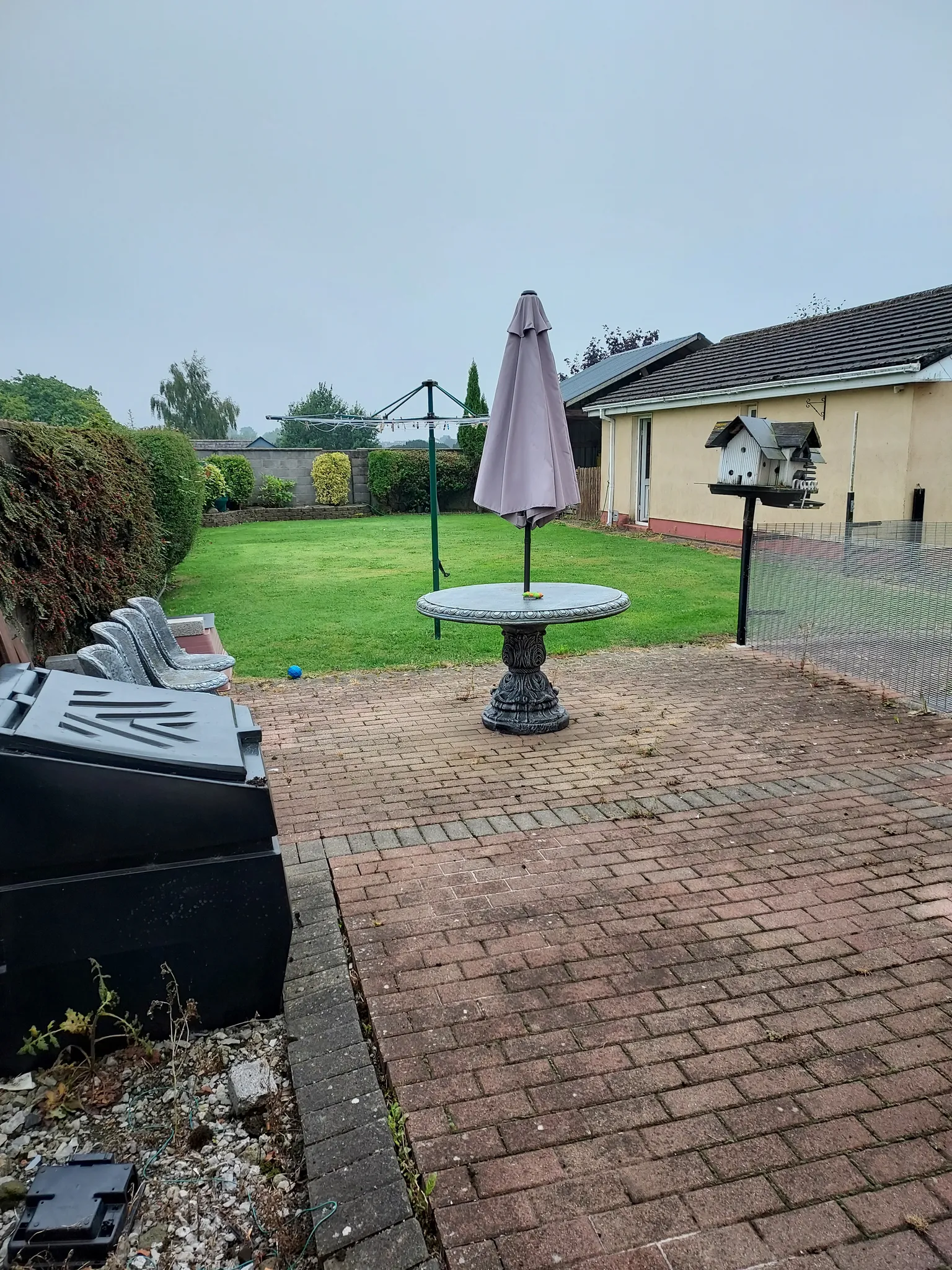Backyard patio and lawn with a round table and closed umbrella, rotary clothesline, birdhouse on a post, and a single-story building at the right