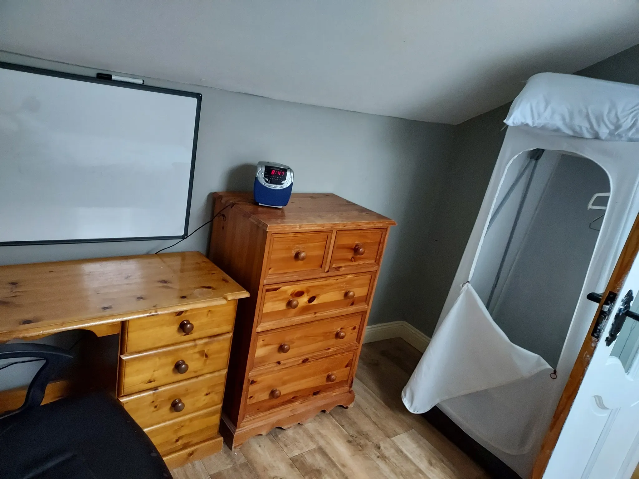 Workspace with a wooden desk and matching tall chest of drawers, wall-mounted whiteboard, office chair, and a hanging garment organizer.