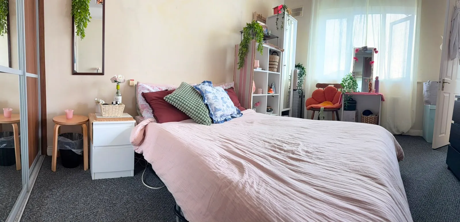 Bedroom with a double bed covered by a pink duvet, several pillows, a bedside table and stool, mirrored wardrobe, shelving unit, orange upholstered chair and a large window with sheer curtains.
